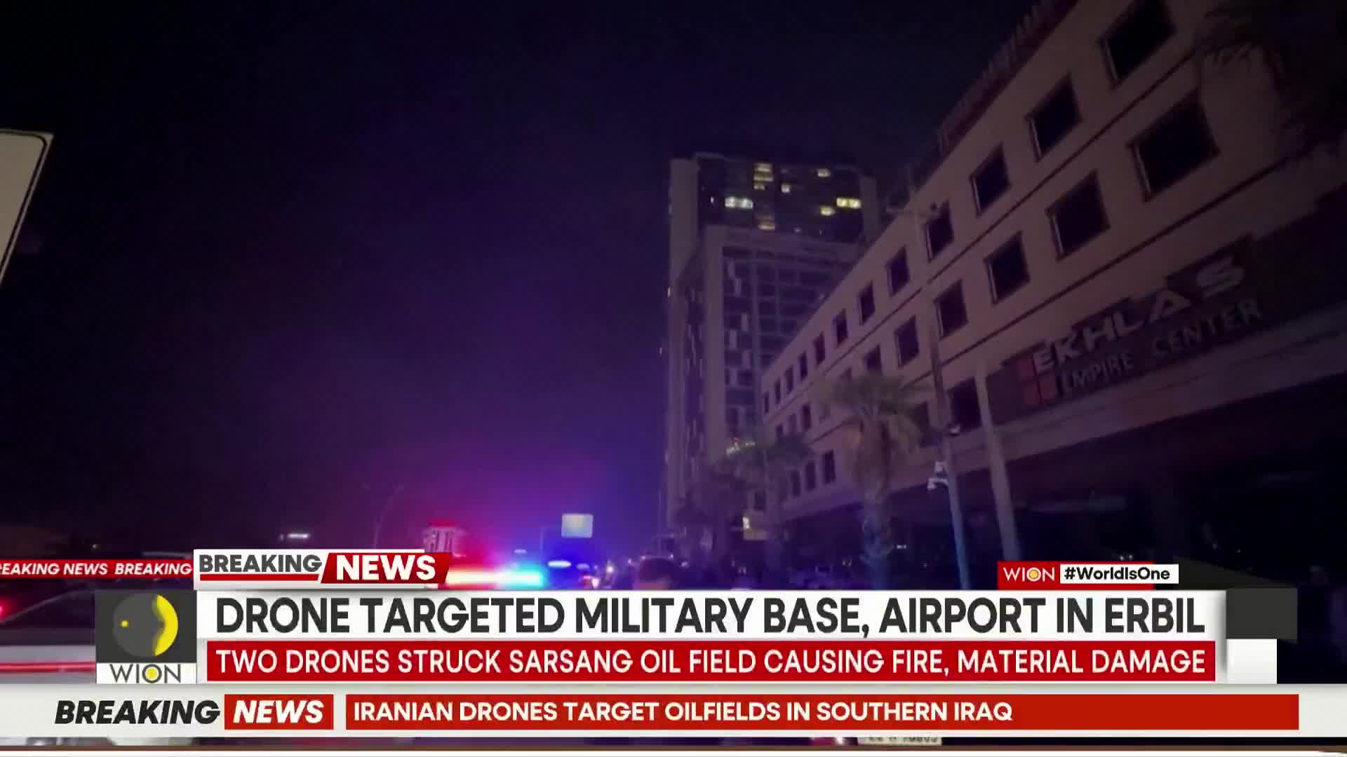 Emergency lights flash red and blue down the street, illuminating a tall building. A news ticker announces drone strikes in Erbil, Iraq.