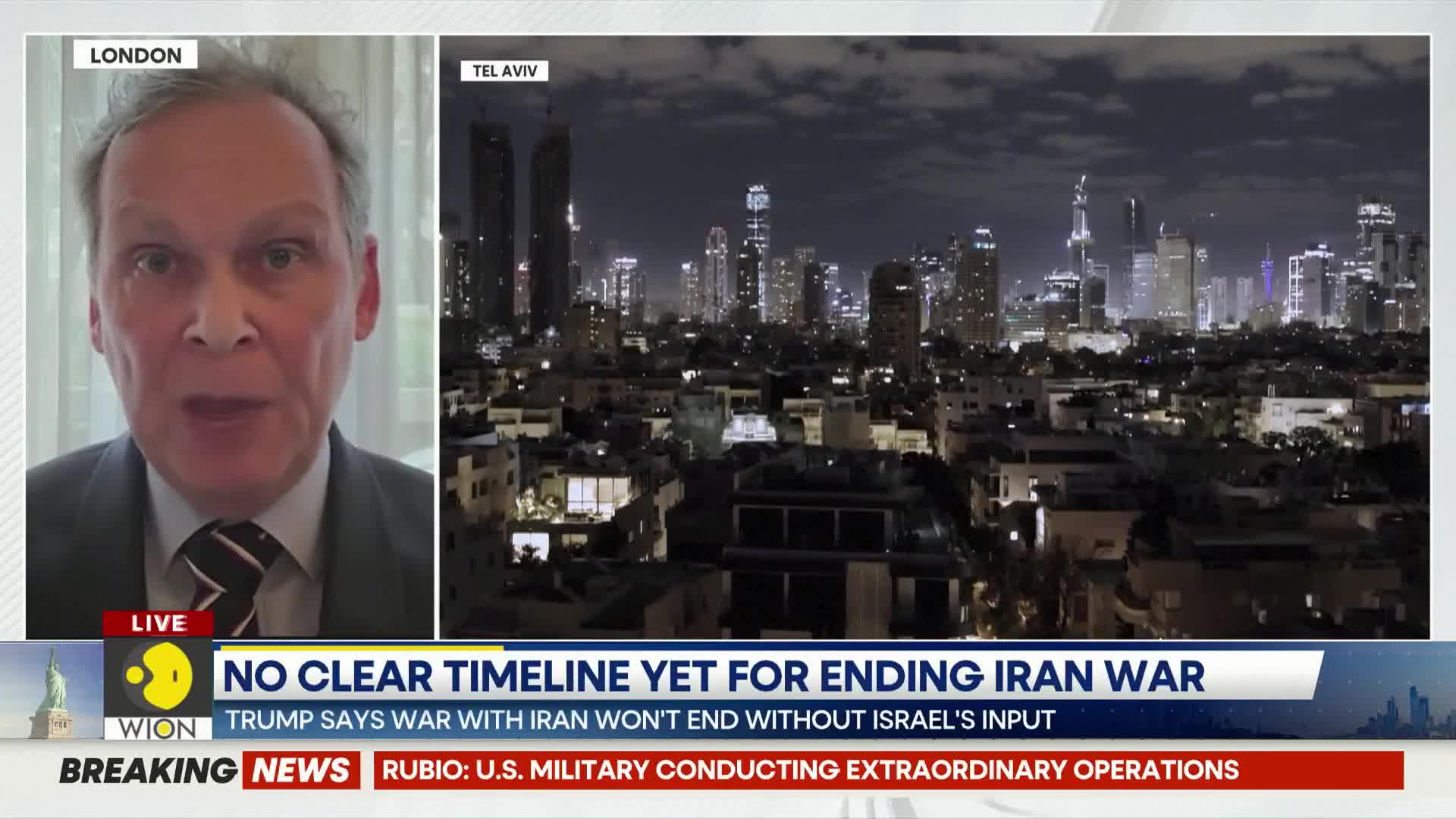 A man speaks from London, his face filling the left side of the screen. Behind him, the Tel Aviv skyline glows with city lights under a dark sky.