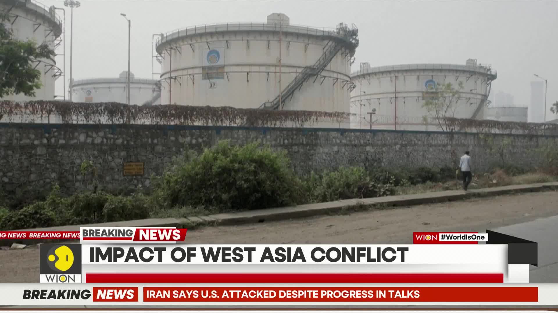 A lone man walks past large storage tanks, their surfaces streaked with grime. A news ticker scrolls by, reporting on the impact of the West Asia conflict.