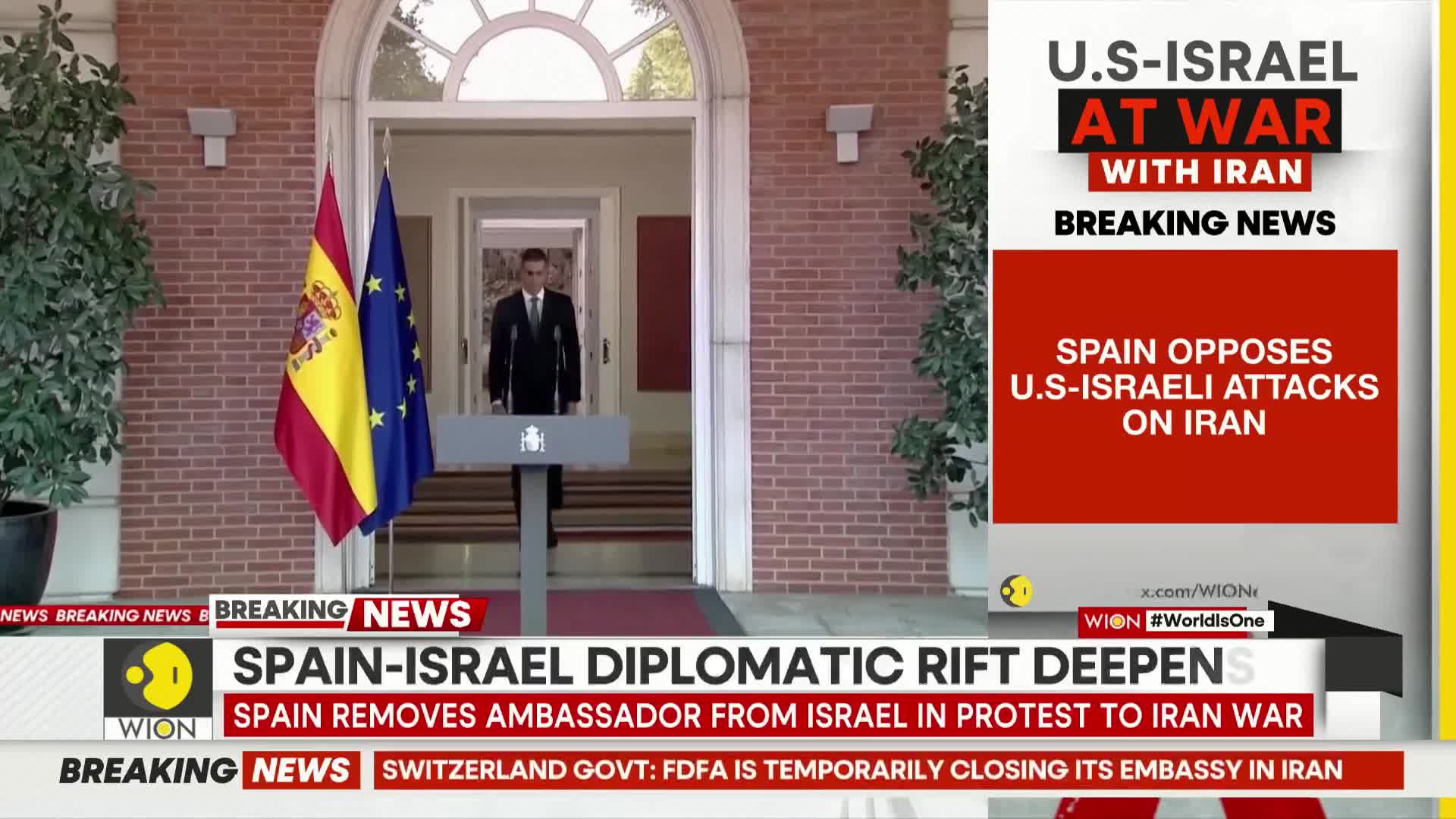 A man in a dark suit stands behind a podium, flanked by the Spanish and EU flags. Breaking news tickers scroll across the bottom of the screen, announcing a diplomatic rift between Spain and Israel over attacks on Iran.