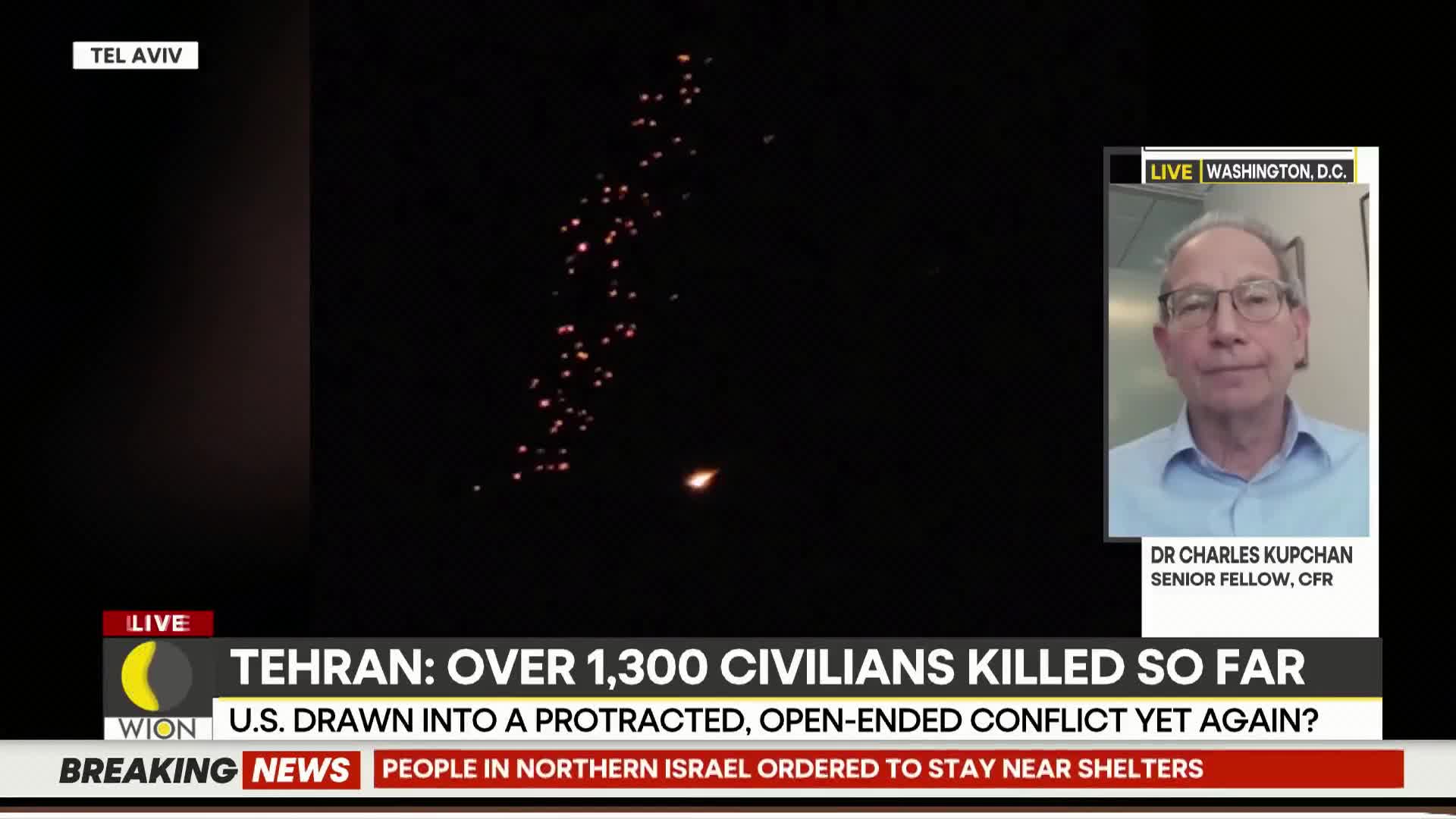 A cluster of bright orange lights arcs across the dark sky, with a single, larger light burning below them. A man in a light blue shirt speaks from Washington D.C. on a screen showing a news report from WION about events in Tehran.