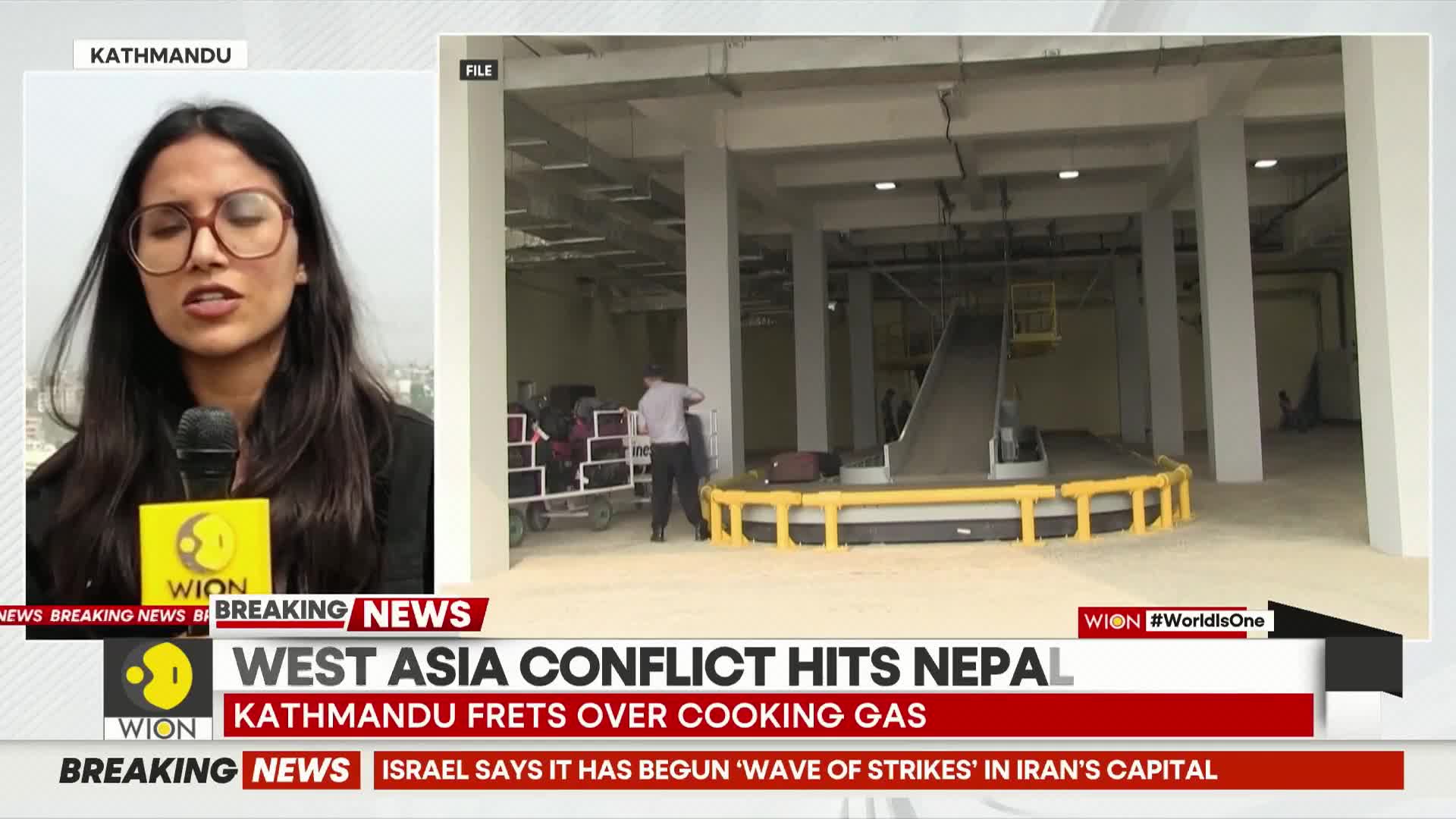 Bags are being loaded onto a conveyor belt in a large, industrial-looking space. A man in a light shirt and dark pants is guiding a cart piled high with luggage towards the belt.