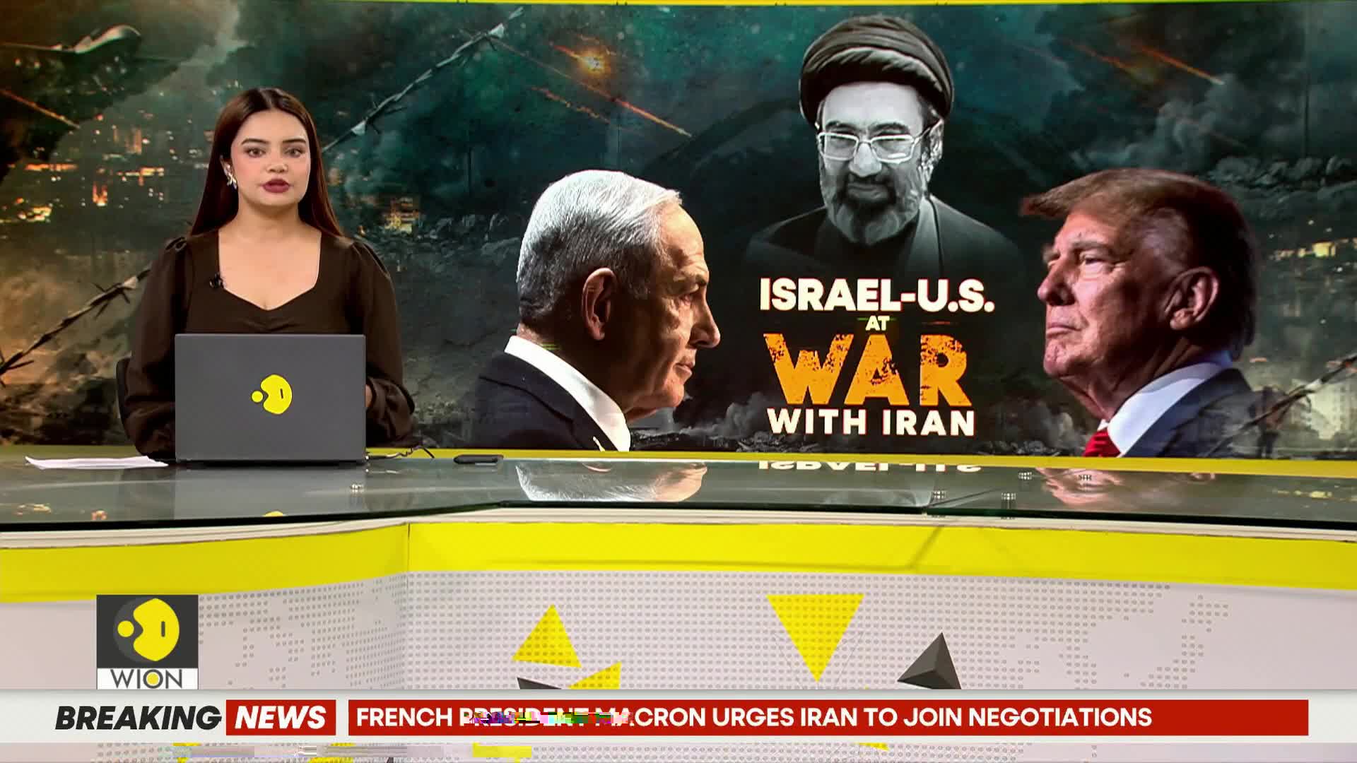 A news anchor in a dark top sits at a desk with a laptop. Behind her, a screen displays portraits of political figures and the headline "ISRAEL-U.S. AT WAR WITH IRAN." A ticker at the bottom of the screen reads "BREAKING NEWS FRENCH PRESIDENT MACRON URGES IRAN TO JOIN NEGOTIATIONS."