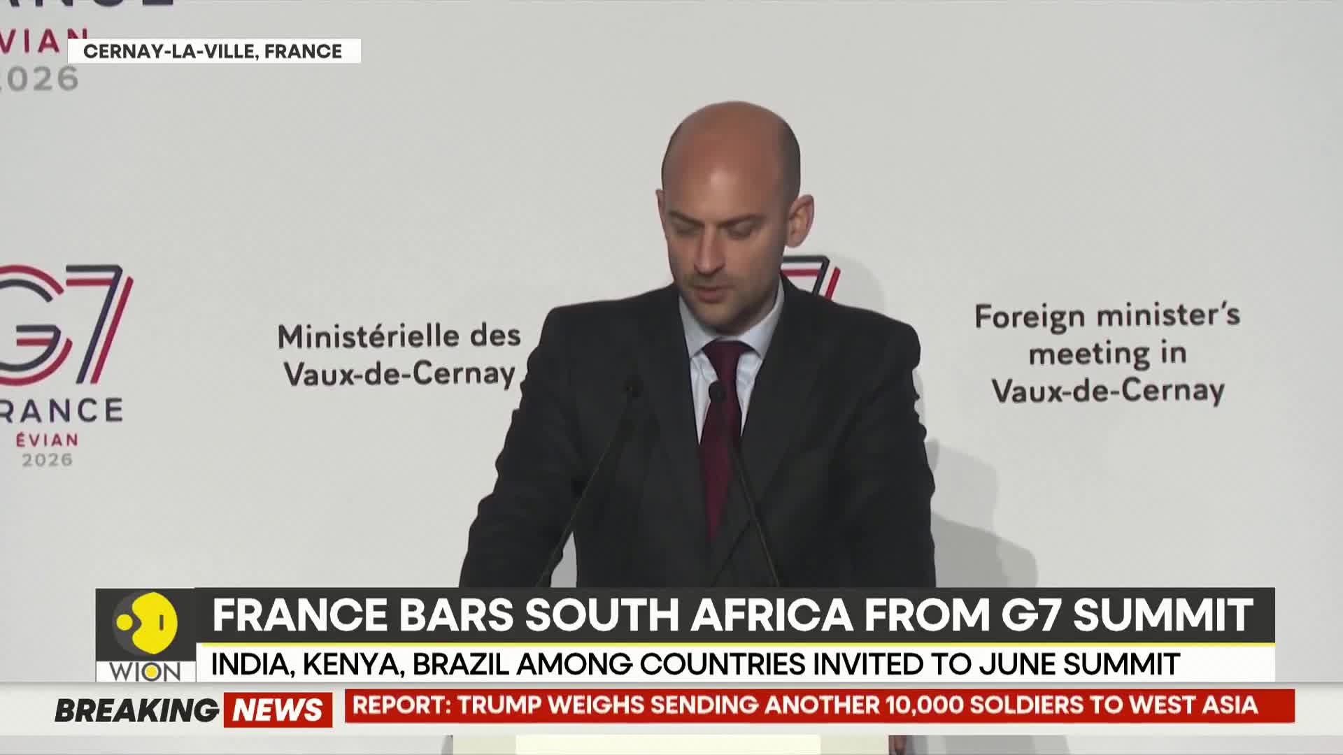 A man in a suit stands at a podium, reading from papers. A news ticker at the bottom of the screen reports on France barring South Africa from a G7 summit, with India, Kenya, and Brazil invited.