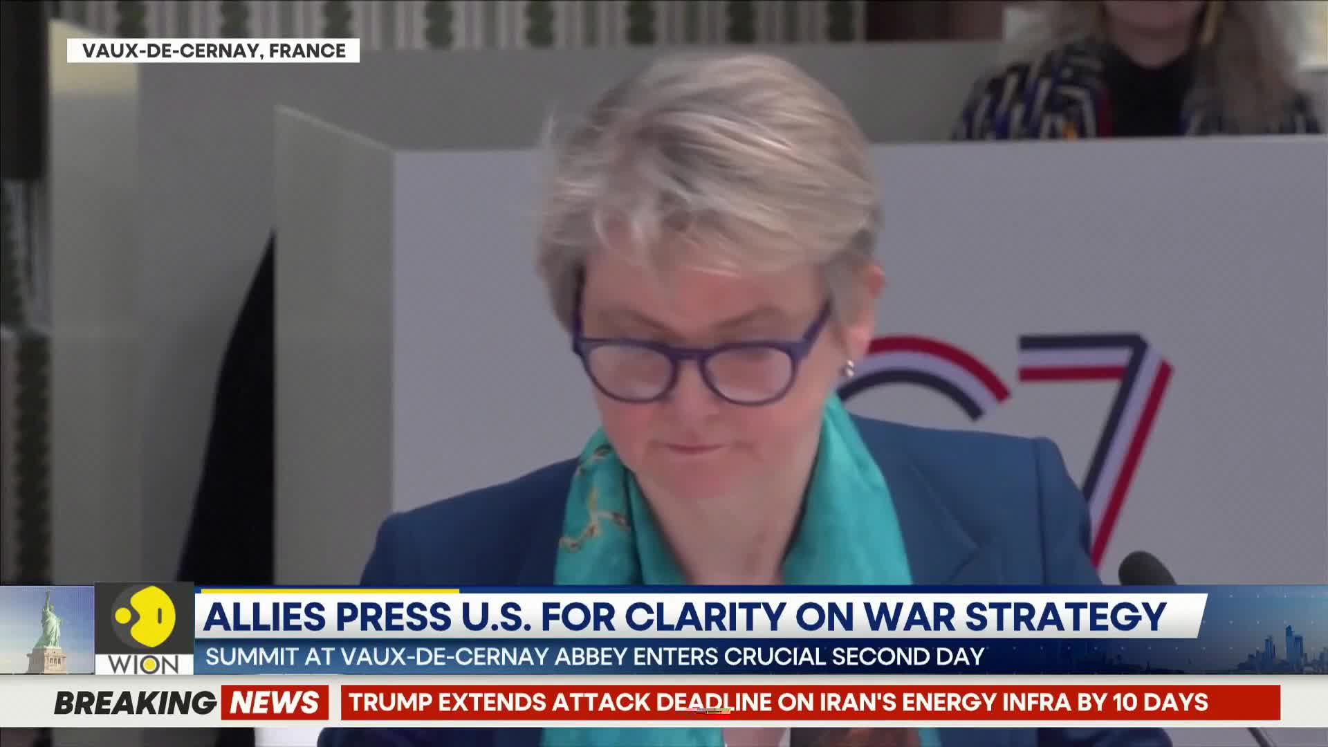 A woman in a blue blazer and teal scarf leans forward, her glasses perched on her nose as she reads. A WION news ticker scrolls at the bottom, reporting on a summit in France and breaking news about Iran.