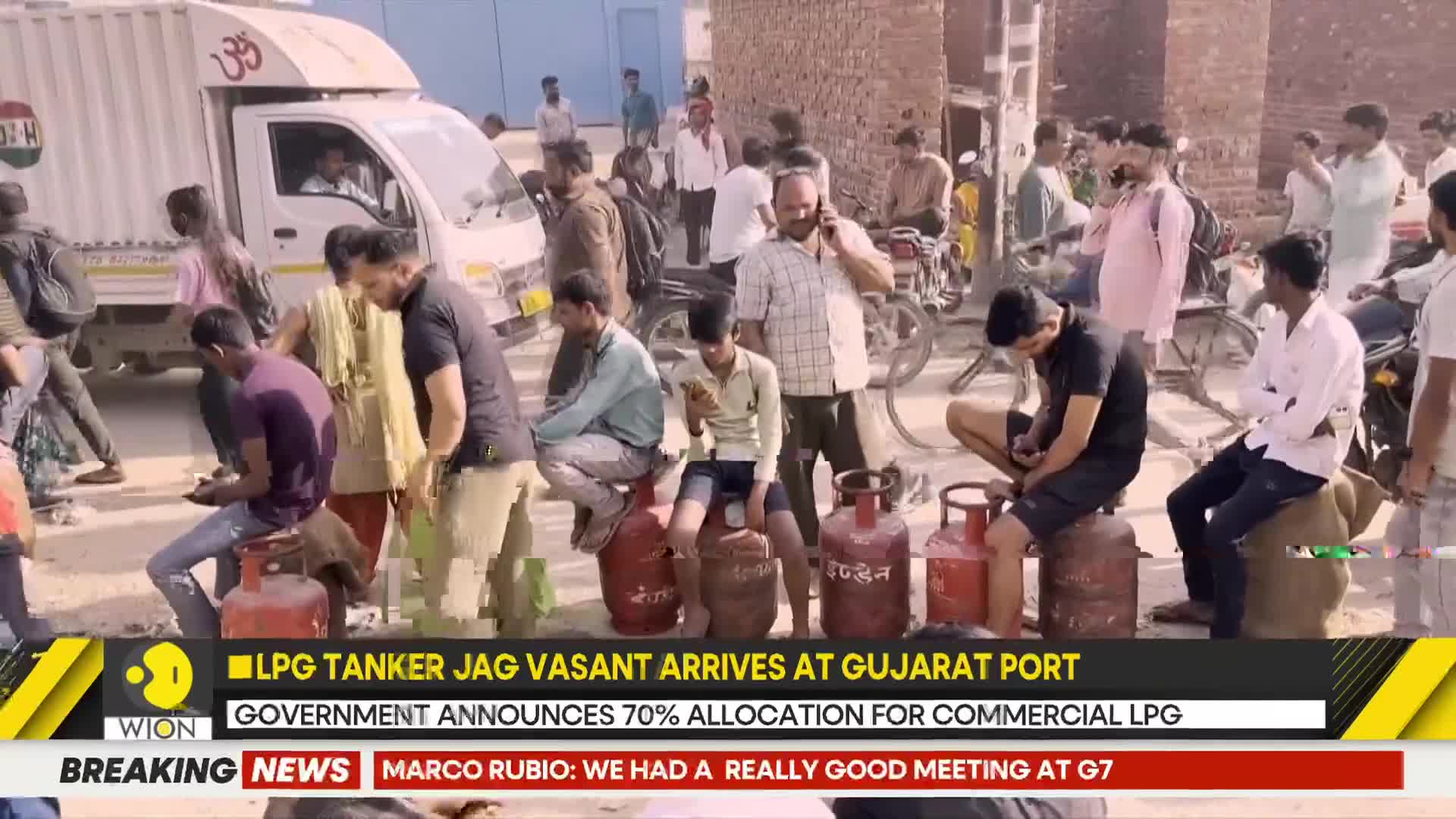 A white truck idles as men sit on red LPG cylinders, some looking at their phones. The scene unfolds under a bright sky, with a news ticker mentioning an LPG tanker's arrival at a Gujarat port.
