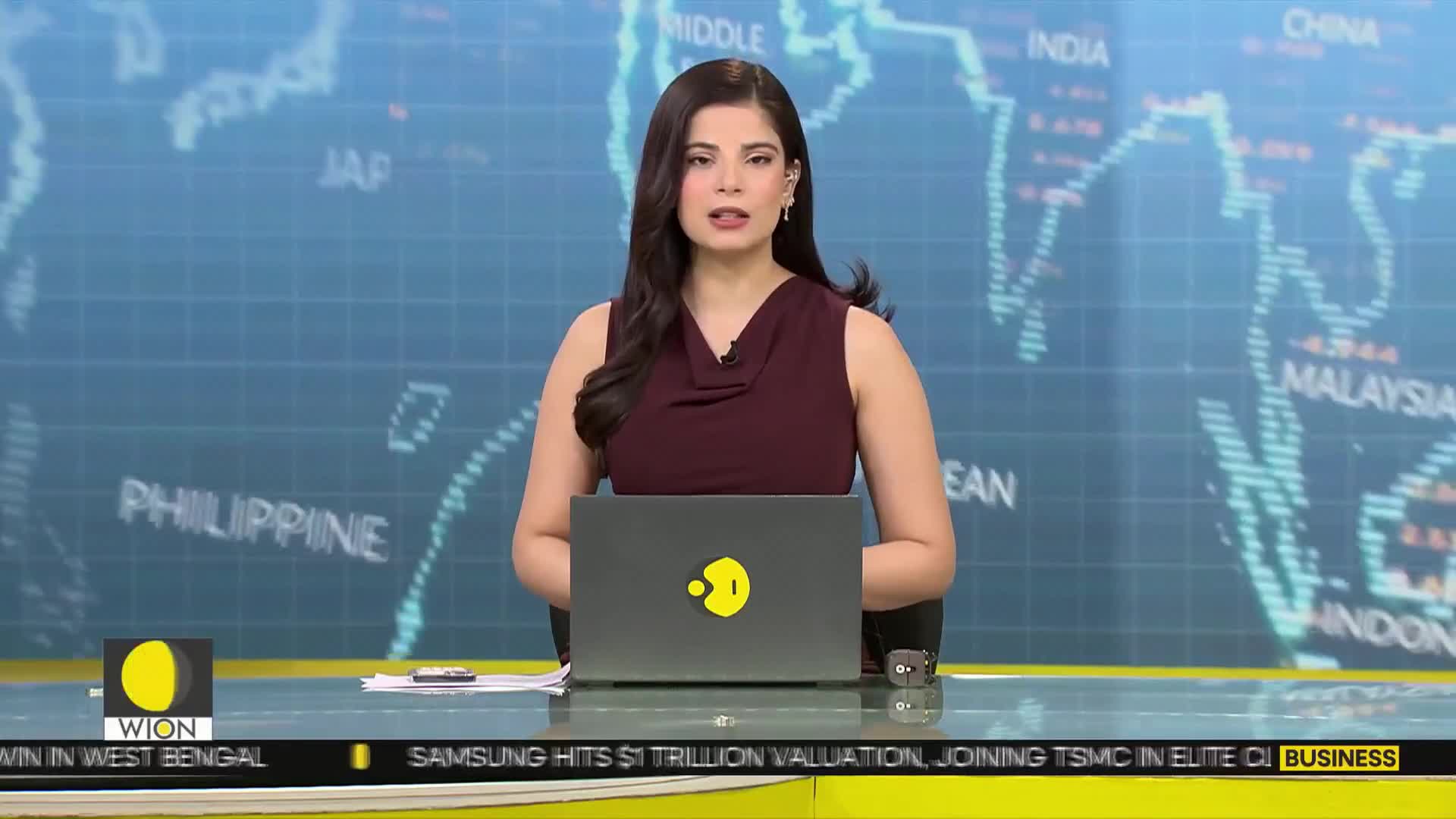 A woman in a maroon dress speaks from behind a desk, a laptop open before her. Behind her, a digital map displays country names like India and Malaysia.
