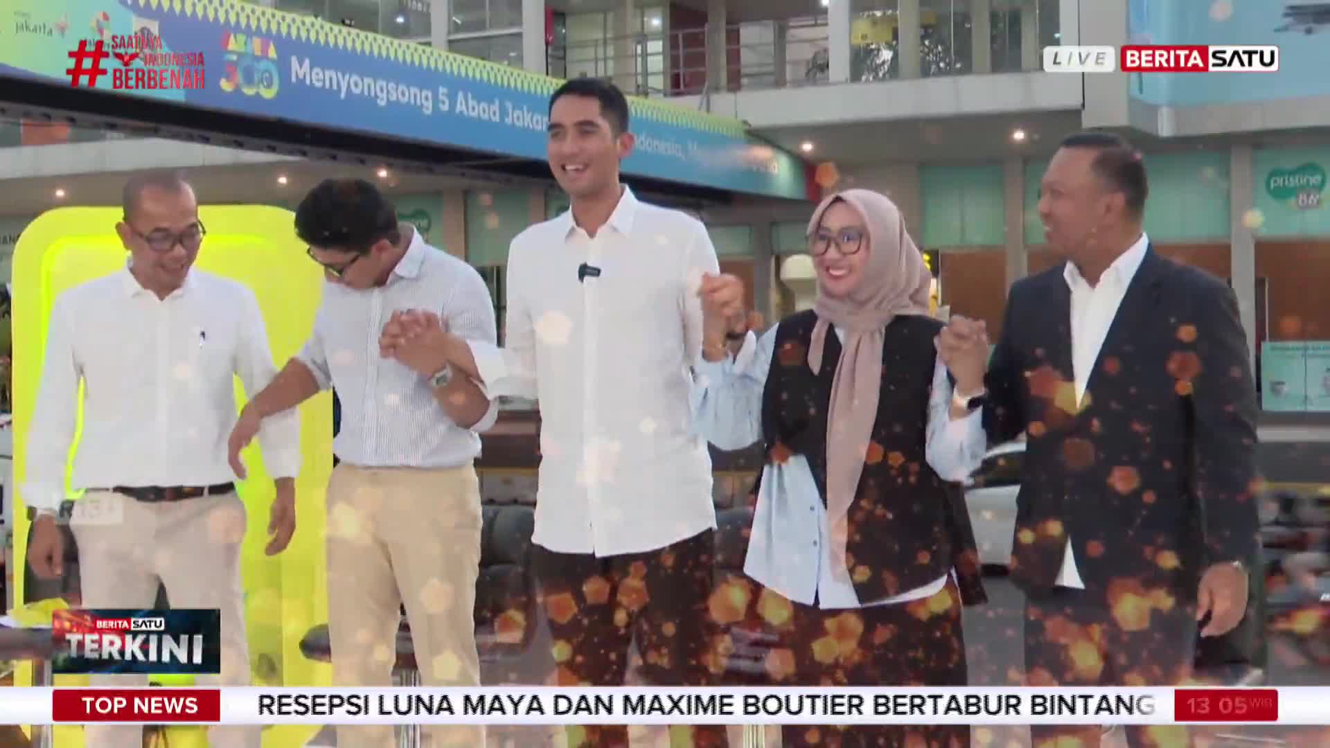 Five people, holding hands, stand on a stage with a "BeritaSatu" logo visible. The backdrop advertises an event for Jakarta's 5th anniversary.
