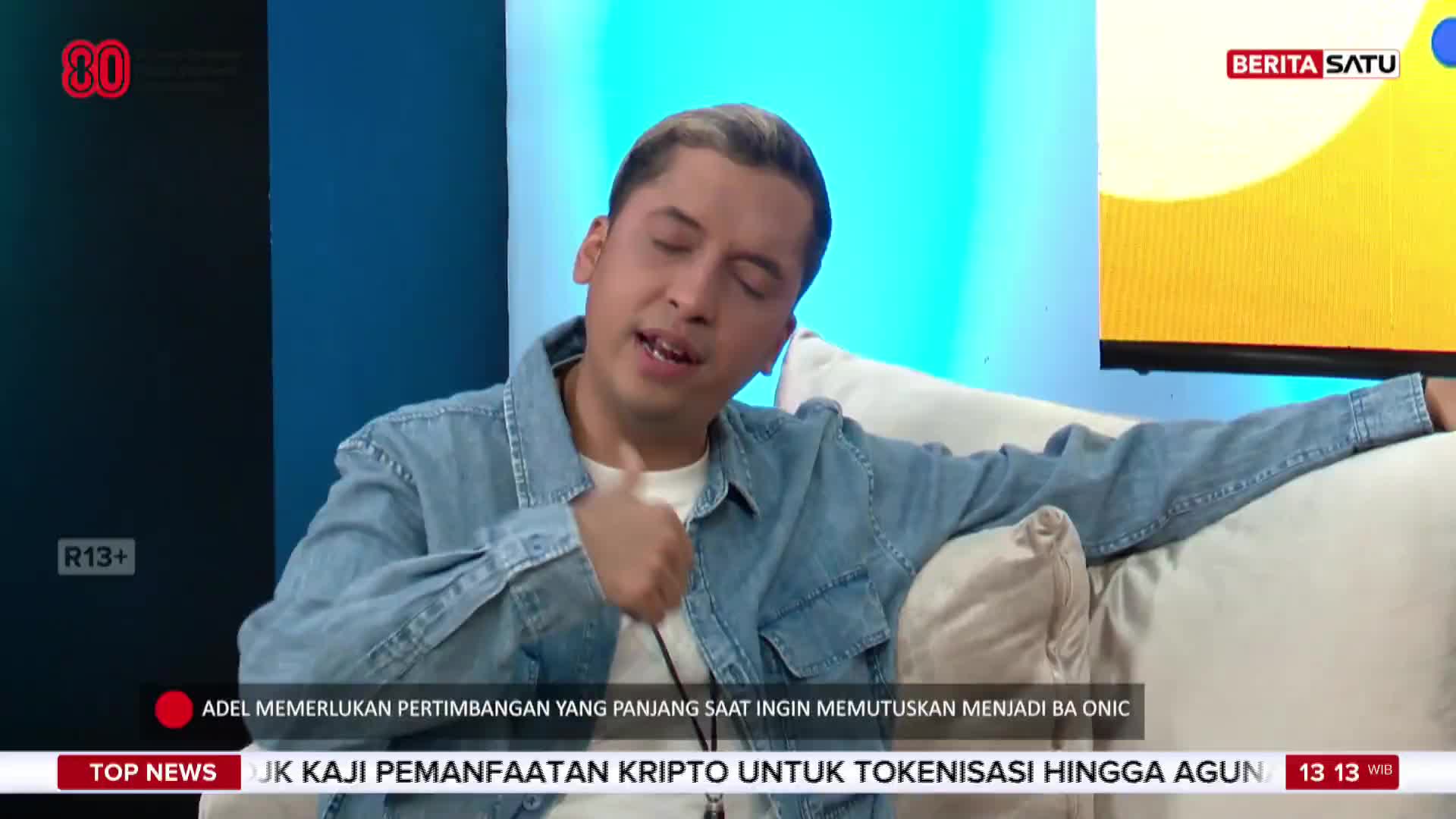 A man in a denim jacket gestures with his right hand while speaking on a BeritaSatu English broadcast. The bottom of the screen displays news headlines in Indonesian, including a mention of crypto.
