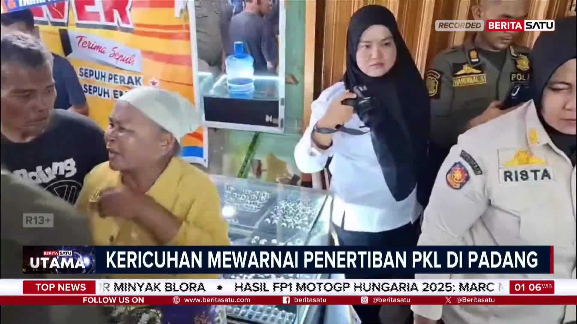 A woman in a yellow shirt gestures emphatically, speaking to someone off-camera as a news crew from BeritaSatu English films the scene. Behind her, a display case of jewelry sits in a shop, and a woman in a hijab looks on, holding a camera.
