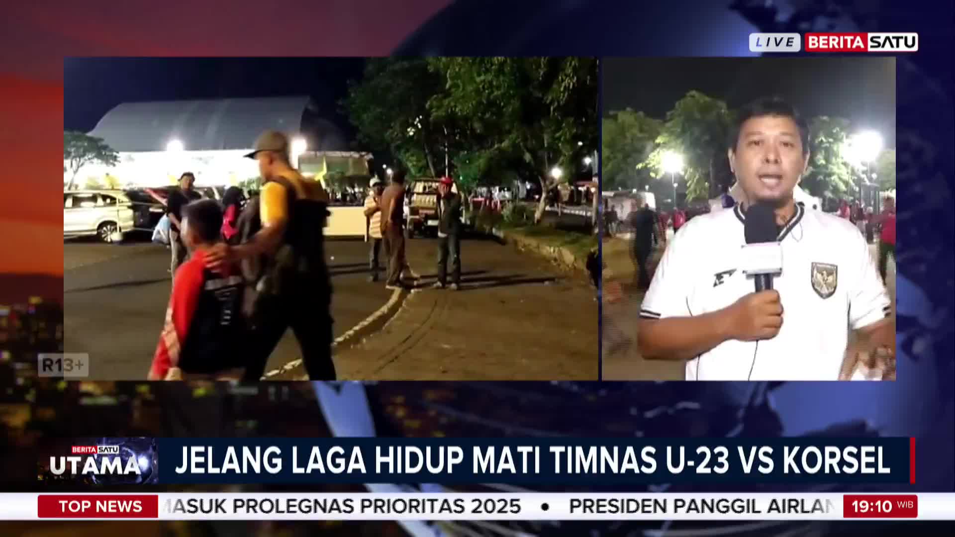 A BeritaSatu English reporter, with the Indonesian flag on his shirt, speaks into a microphone. In the background, people mill about on a dark street near a large building, likely before the U-23 match against South Korea.
