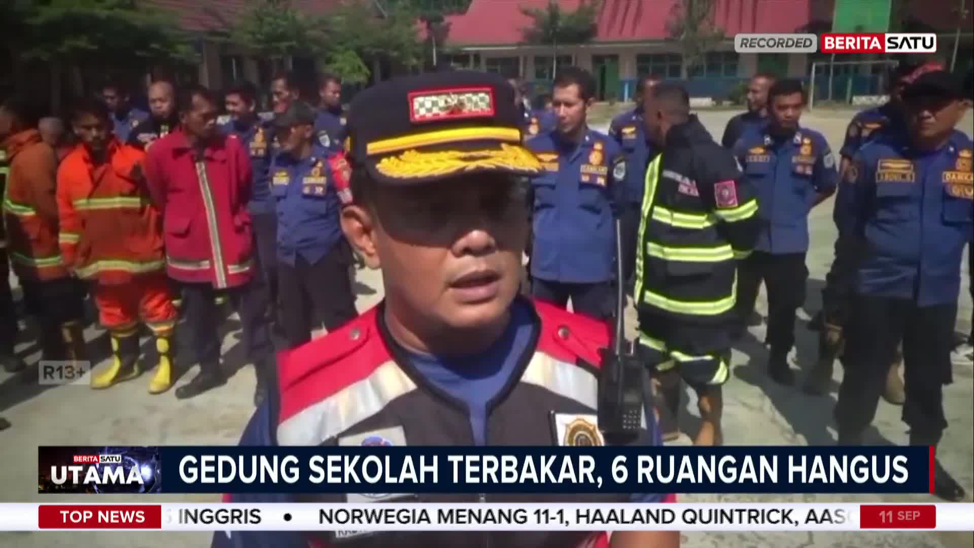 A man in a red and white vest speaks directly to the camera, a group of firefighters standing behind him. The BeritaSatu English headline reports a school building fire in Indonesia, with six rooms destroyed.
