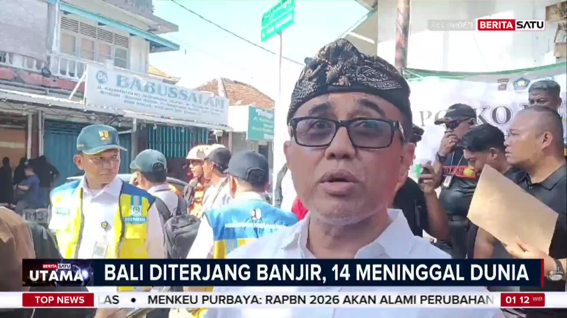 A man in glasses and a traditional Balinese head covering speaks directly into a camera, likely for BeritaSatu English. Behind him, a crowd of people gathers near a building, the backdrop indicating a scene from Indonesia.
