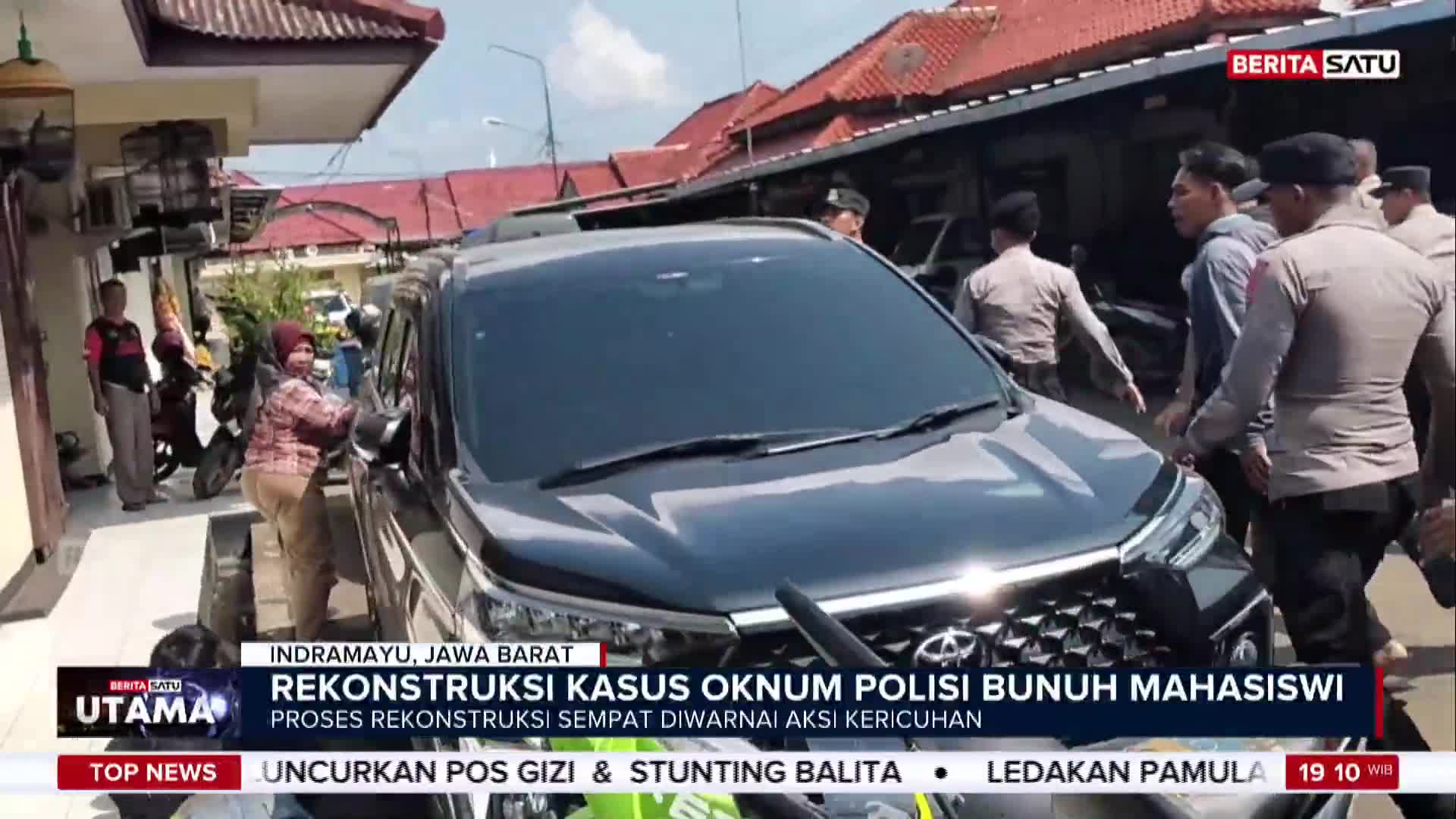A black car is surrounded by several men, some in uniform. A woman in a headscarf stands near the car, seemingly interacting with someone inside.
