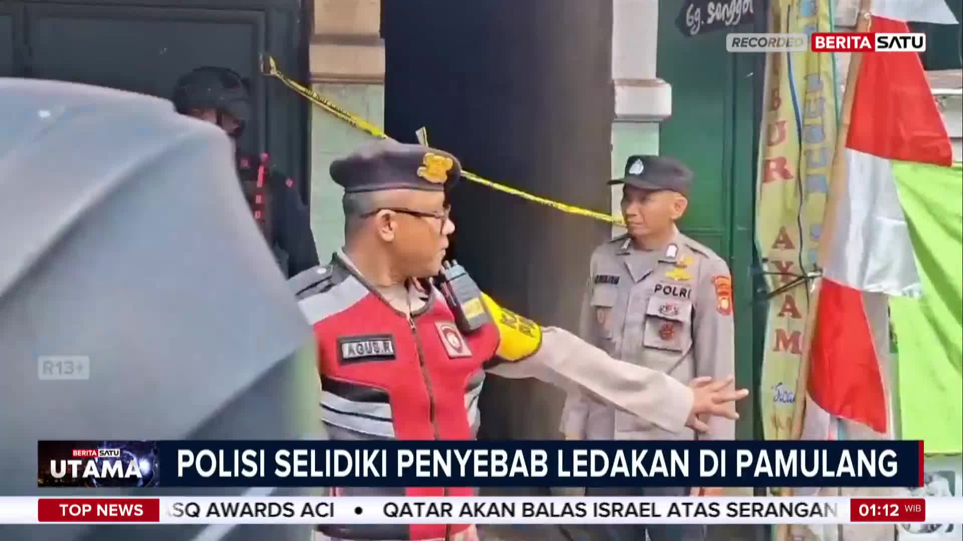 A police officer in a red vest and glasses gestures towards a building entrance cordoned off with yellow tape. The BeritaSatu English news banner reports on a police investigation into an explosion in Pamulang, Indonesia.
