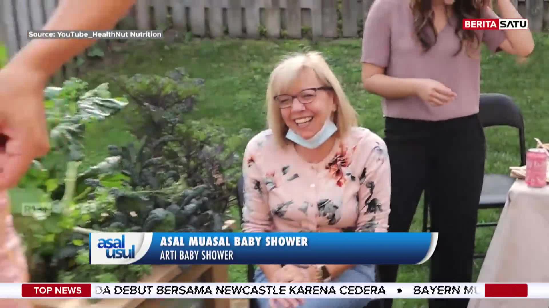 A woman in a floral cardigan laughs, her face mask pulled down, as she sits outside. The BeritaSatu English logo and a banner about a baby shower are visible on the screen.
