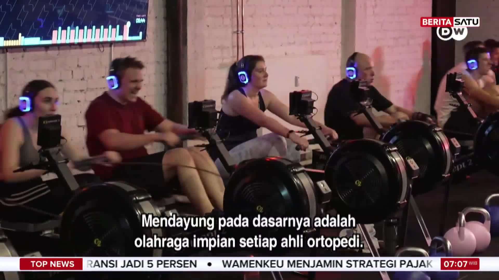 People wearing headphones are pulling on rowing machines, their faces focused as they slide back and forth. The scene appears to be part of a BeritaSatu English news segment from Indonesia, with text about orthopedic health.
