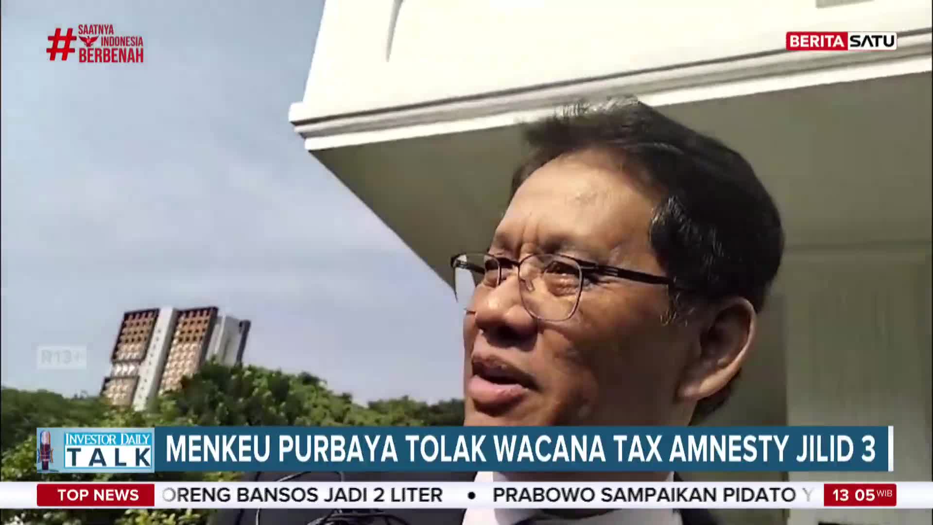 A man in glasses is speaking, likely for BeritaSatu English, with a headline about tax amnesty scrolling beneath him. Behind him, a building's white facade and some trees are visible under a bright sky in Indonesia.
