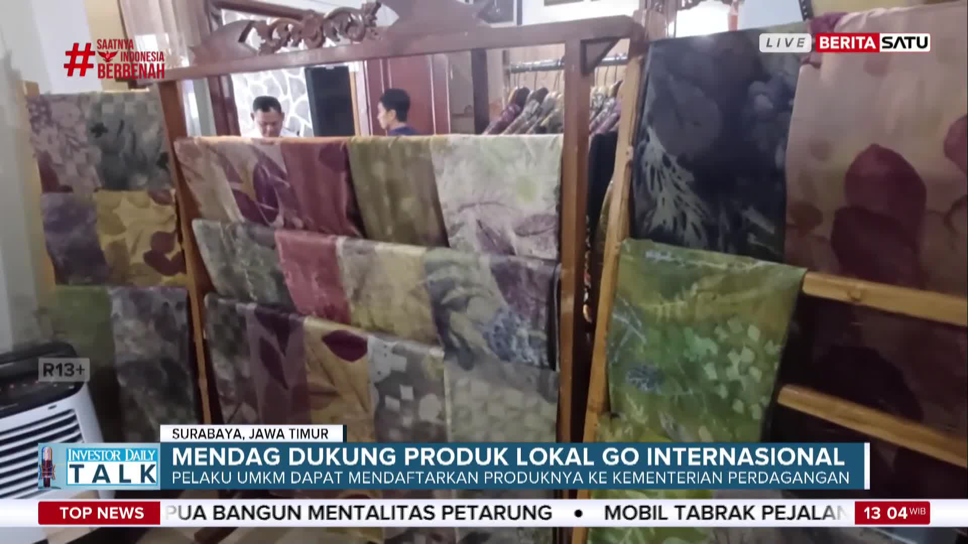 Colorful batik textiles are neatly displayed on a wooden rack, likely for sale. The scene is part of a BeritaSatu English broadcast from Surabaya, Indonesia, discussing local products going international.
