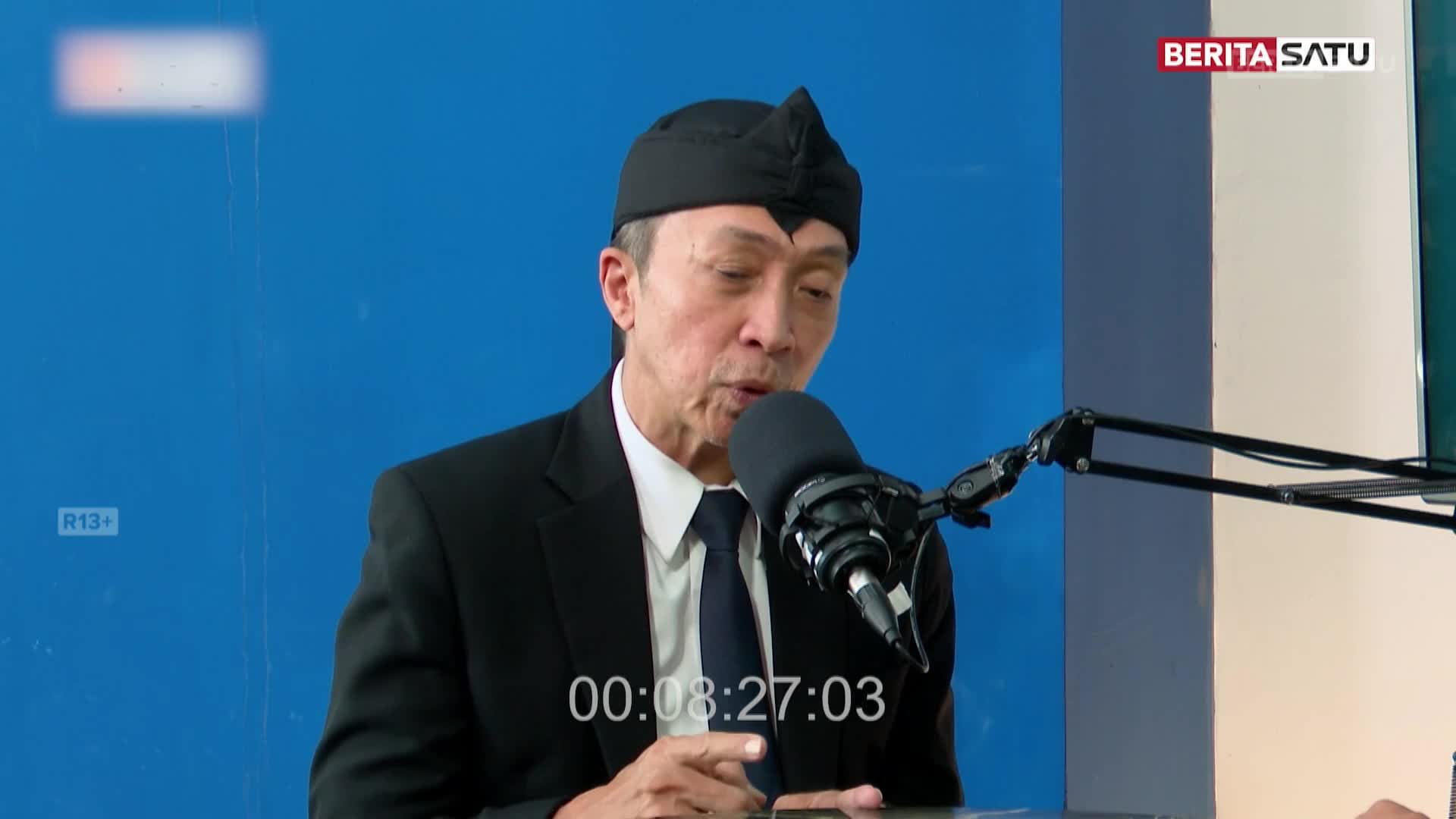 A man in a black suit and traditional headwear speaks into a microphone, likely on BeritaSatu English. The timecode on the screen reads 00:08:27:03.
