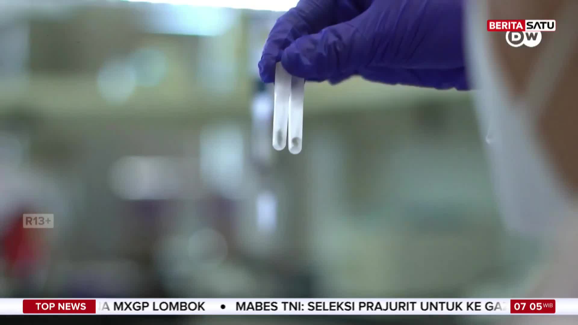 A gloved hand holds two small, clear vials, likely in a lab setting, as seen on BeritaSatu English. The news ticker at the bottom mentions "MXGP Lombok" and "Seleksi Prajurit," indicating a focus on Indonesian news.
