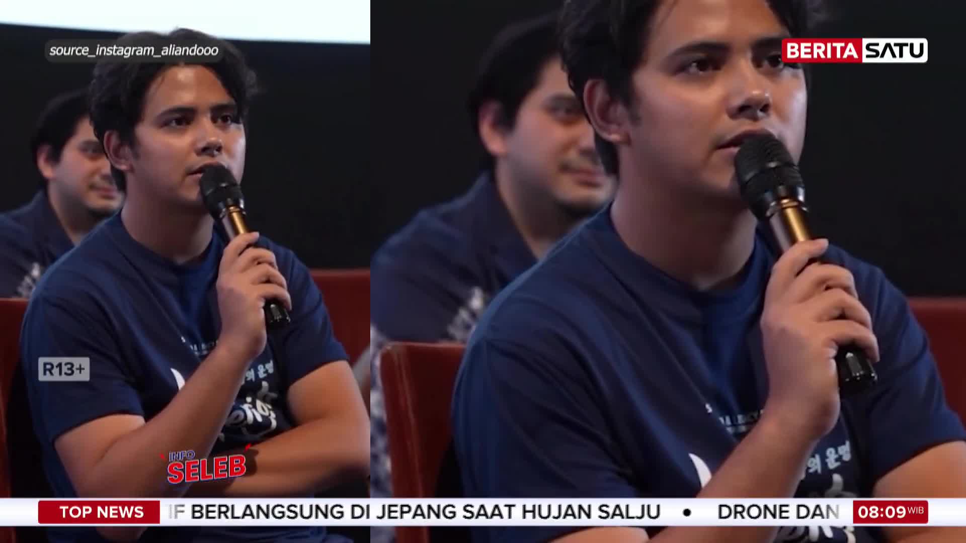 A young man in a dark blue t-shirt holds a microphone to his lips, speaking into it. Behind him, other men in similar shirts sit in chairs, their faces blurred.