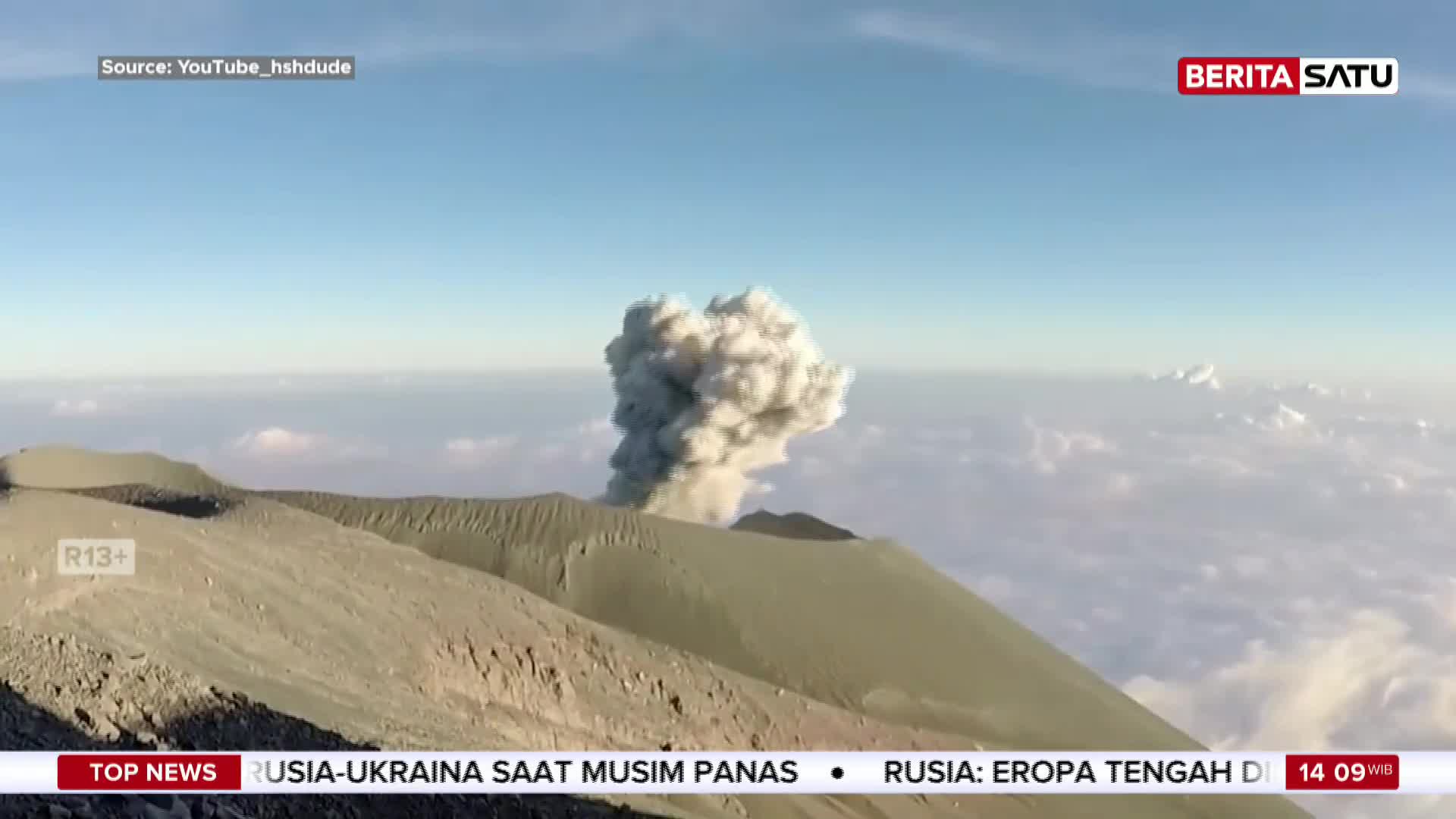 A massive plume of ash erupts from the summit of a volcano, rising high into the clear blue sky. Below, the rugged, sandy slopes of the mountain are visible, with clouds stretching out towards the horizon.