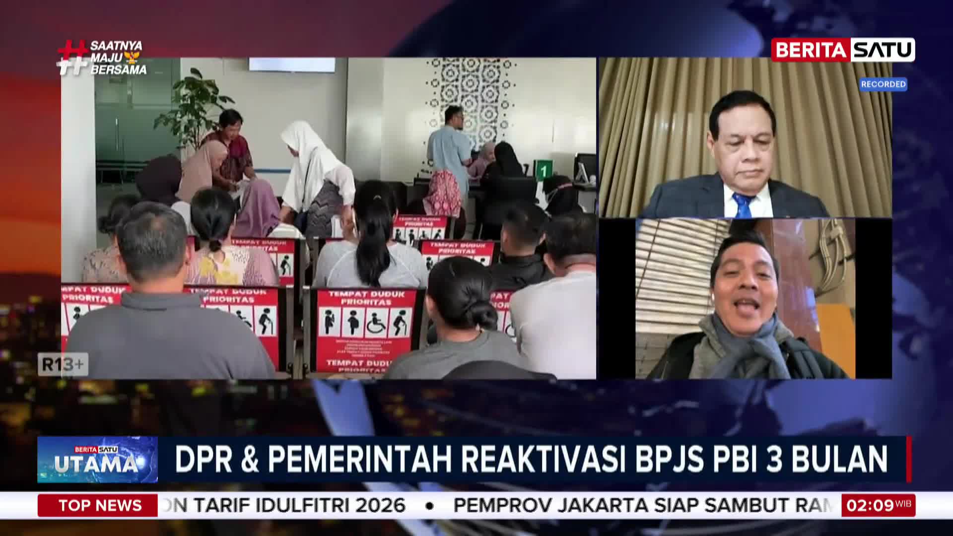 People are waiting in a government office, some seated and others standing near a counter. On a screen, two men are speaking, one in a suit and the other in a scarf.