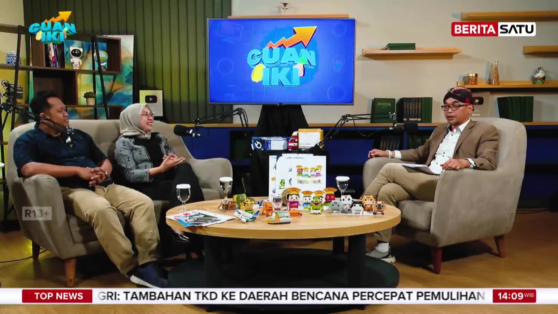 A panel of three people are seated on couches around a coffee table, discussing something as a BeritaSatu English broadcast plays behind them. The screen displays the "Guan Iki" logo, a program title.