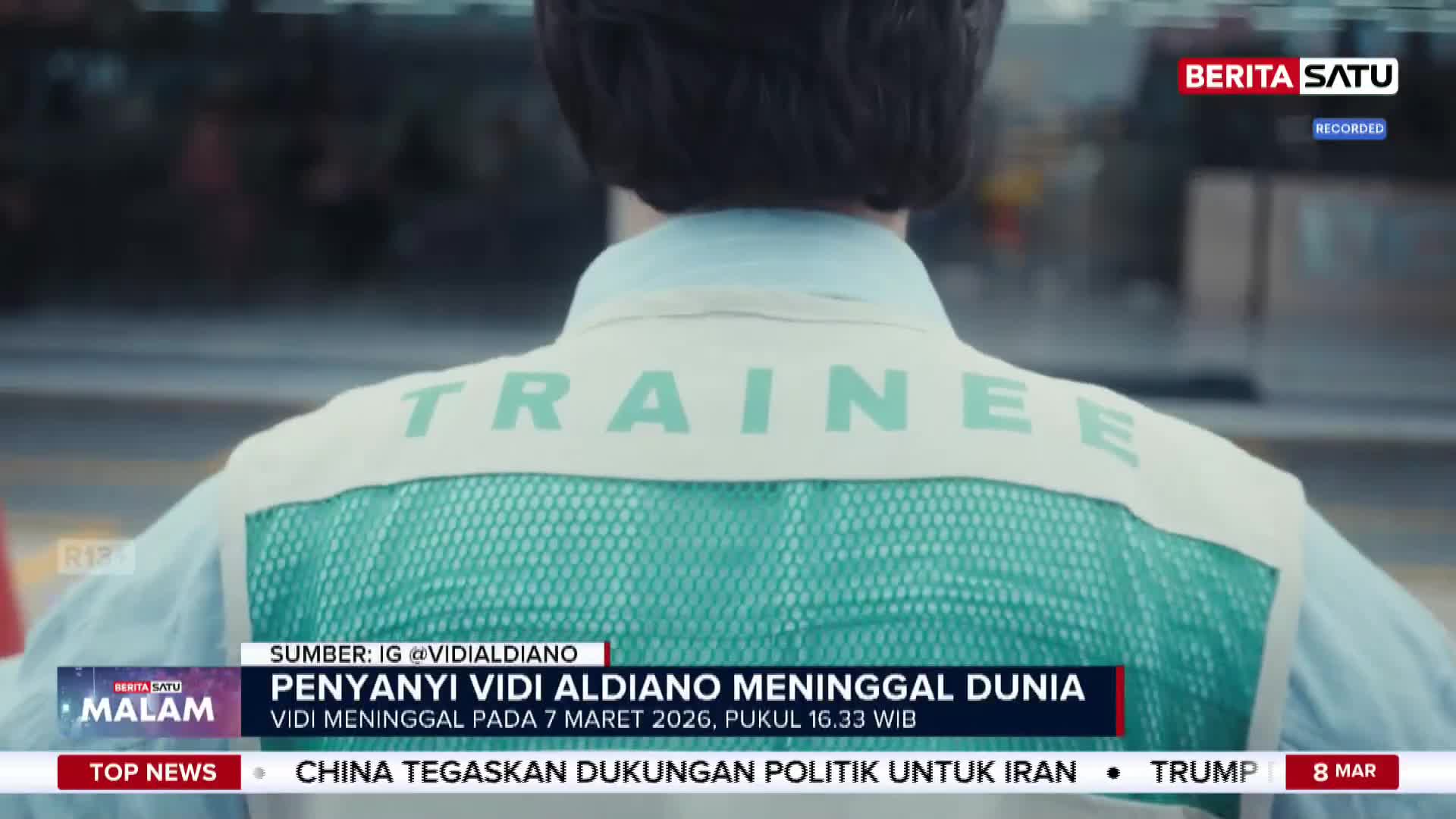 A person wearing a vest with "TRAINEE" printed on the back walks away. A news ticker from Berita Satu English scrolls across the bottom of the screen.