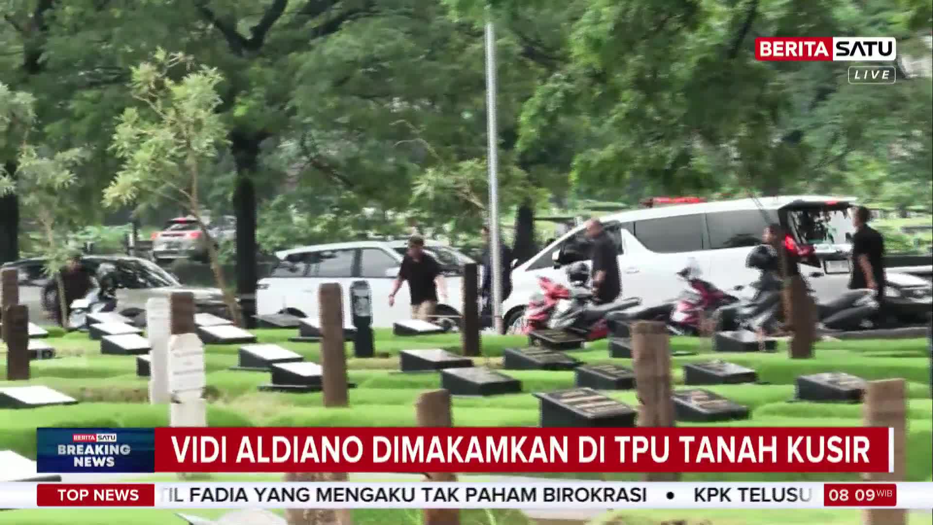 People in dark shirts move between white vans and motorcycles parked among rows of gravestones at TPU Tanah Kusir. A BeritaSatu live broadcast overlays the scene, reporting on Vidi Aldiano's burial.