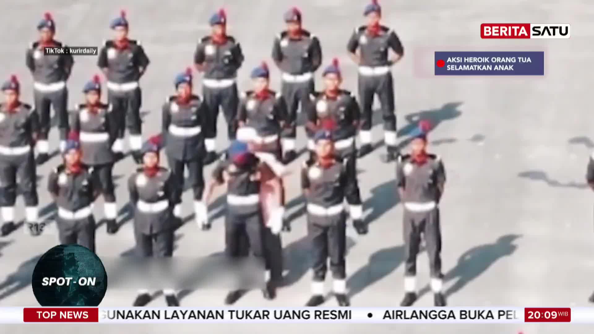 A group of uniformed individuals stand at attention, their dark uniforms accented with white belts and red and blue caps. One person in the center, wearing a pinkish garment, is being assisted by another in uniform. A news ticker at the bottom displays "TOP NEWS" and Indonesian text.