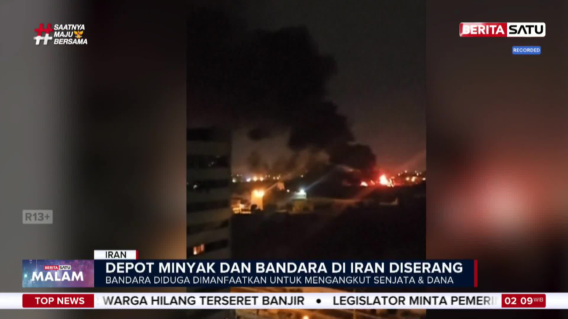 Thick black smoke billows into the night sky over a city. Flames erupt from multiple points on the ground, illuminating the scene with an orange glow.