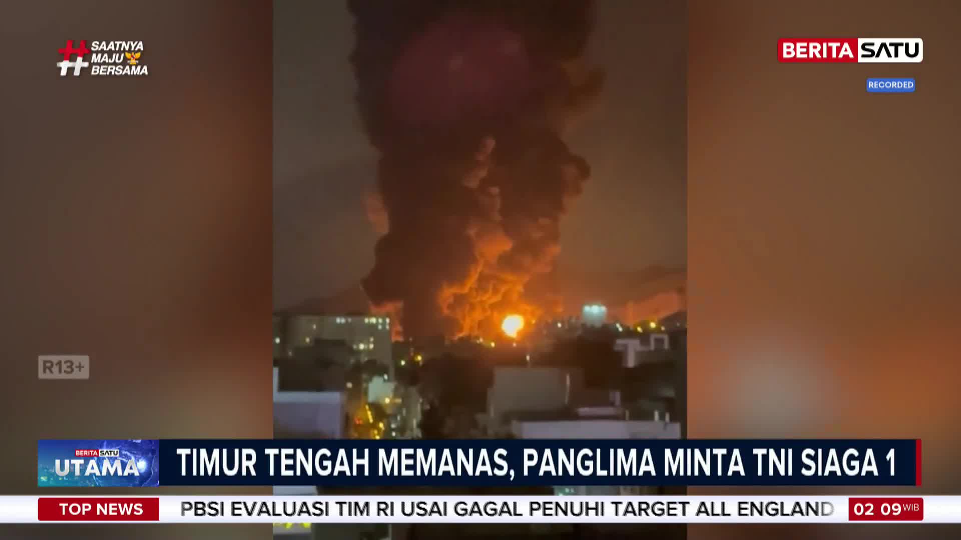 A massive plume of dark smoke billows into the night sky, illuminated by intense orange flames. Below, a city skyline glows with scattered lights.