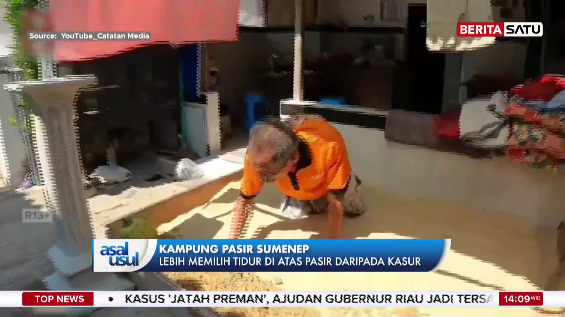 An older man, wearing an orange shirt, crawls on his hands and knees across a sandy surface. He's in front of a building, with what looks like bedding piled up to his right.