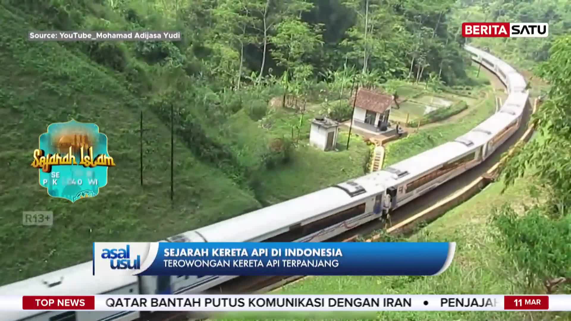 A long, white train snakes its way through a lush, green valley. The train is moving along the tracks, its carriages a sharp contrast to the verdant landscape of Indonesia.