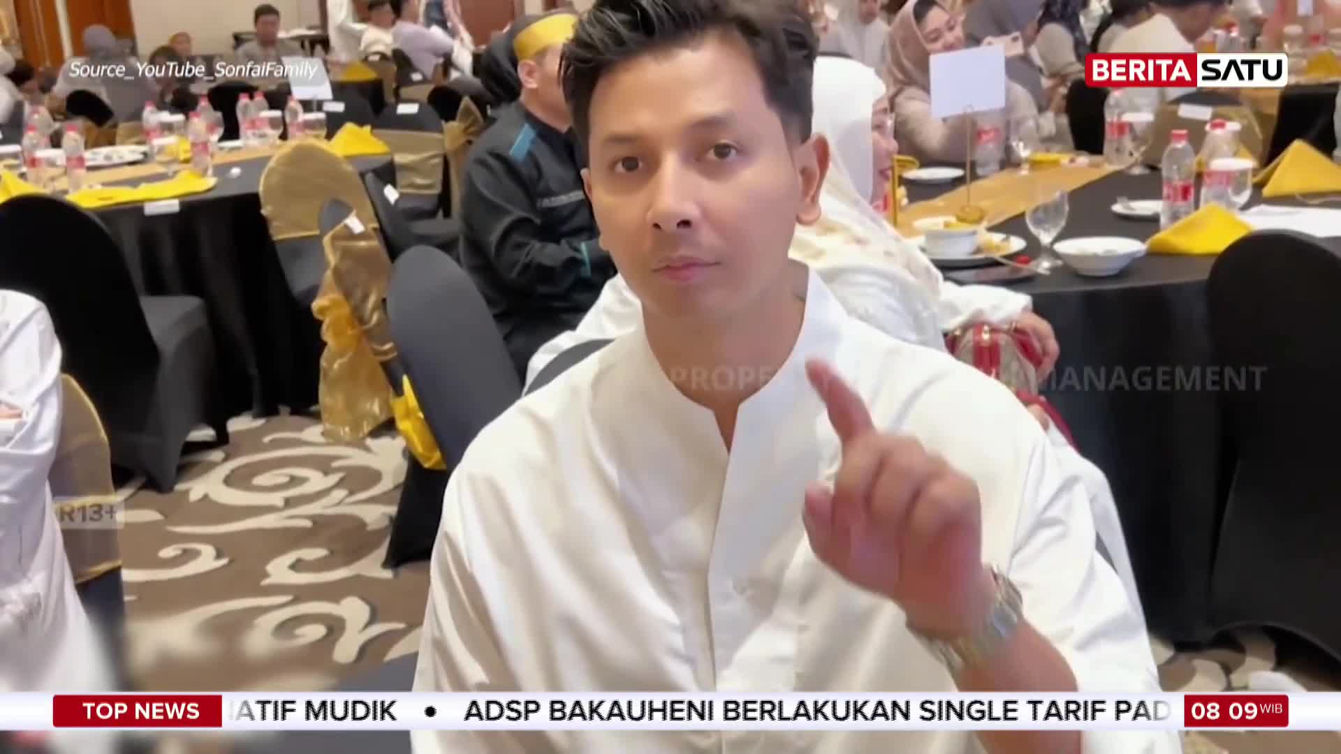 A man in a white shirt gestures with his right hand, his expression serious. Behind him, guests are seated at tables adorned with yellow and black cloths.