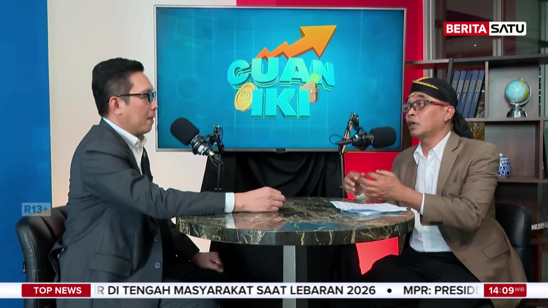 Two men sit across from each other at a table, microphones between them. The man on the right, wearing a traditional Indonesian headscarf, gestures with his hands as he speaks. A screen behind them displays the BeritaSatu logo and the words "CUAN IKI."