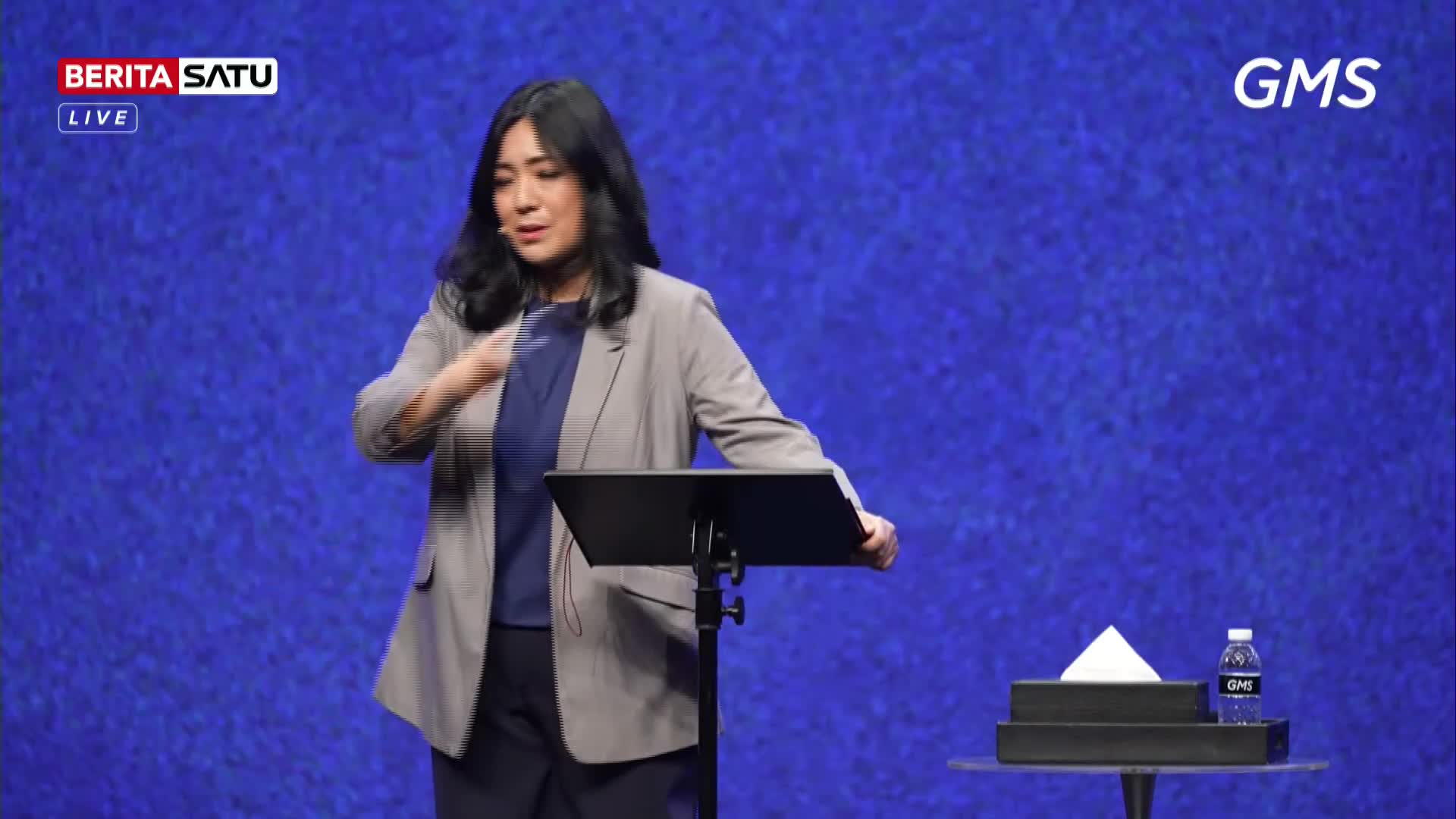 A woman stands at a podium, gesturing with her left hand. A bottle of water and a tissue box sit on a small table to her right.