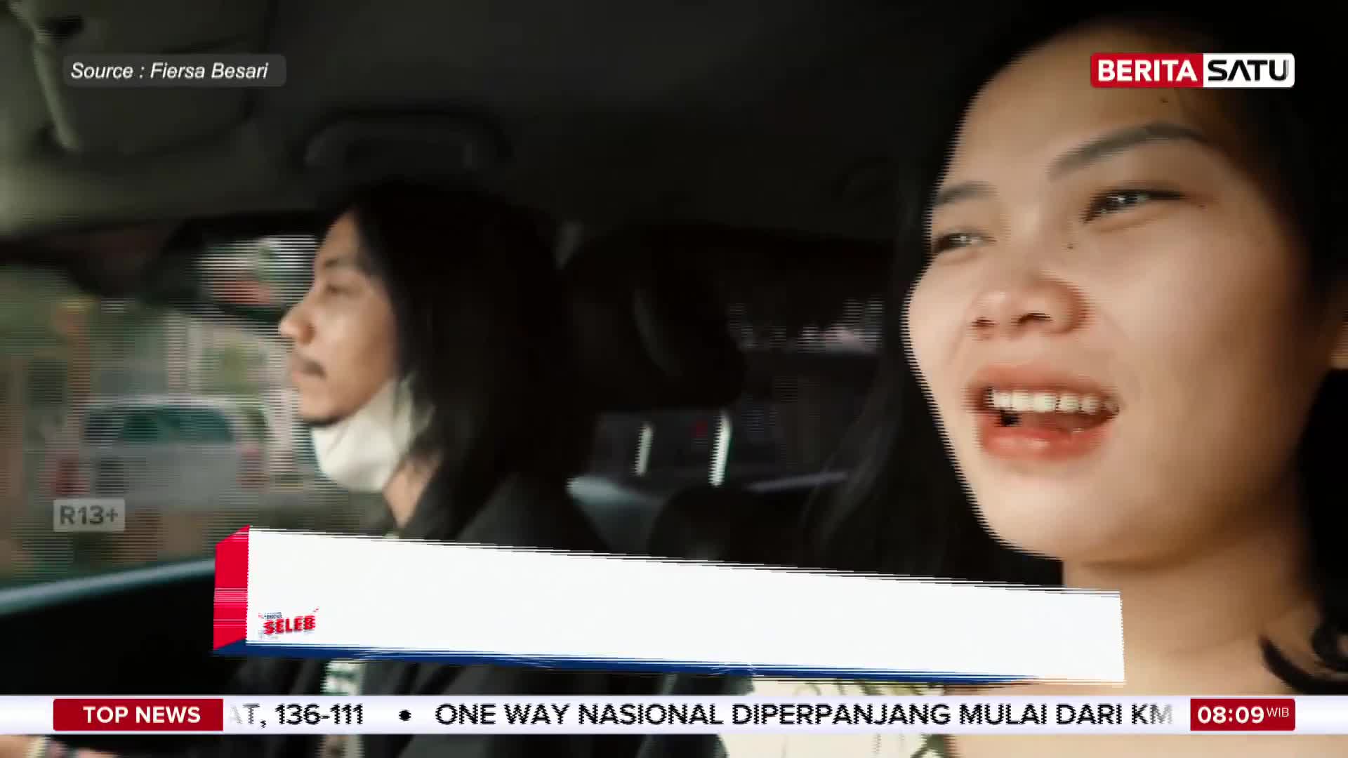 A woman with dark hair laughs animatedly in the passenger seat of a car. Beside her, a man with long hair, wearing a mask, looks ahead as he drives.