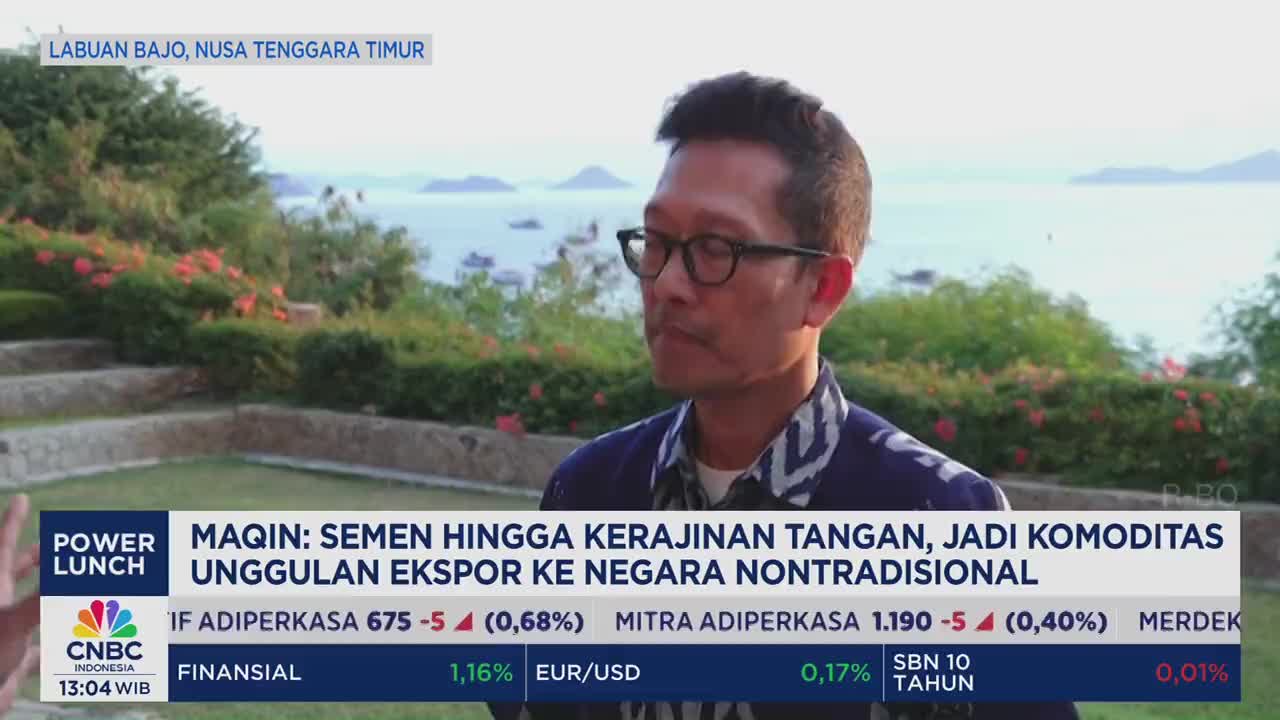 A man in glasses is speaking on CNBC Indonesia, framed against a backdrop of the sea and greenery in Labuan Bajo. The broadcast's lower third displays financial data and the headline "MAQIN: SEMEN HINGGA KERAJINAN TANGAN, JADI KOMODITAS UNGGULAN EKSPOR KE NEGARA NONTRADISIONAL."

