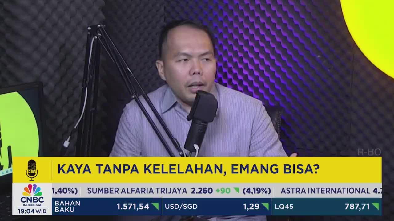 A man in a light-colored shirt speaks into a microphone, likely on a CNBC Indonesia program. The screen displays financial data, with a question about wealth and effort in Indonesian.
