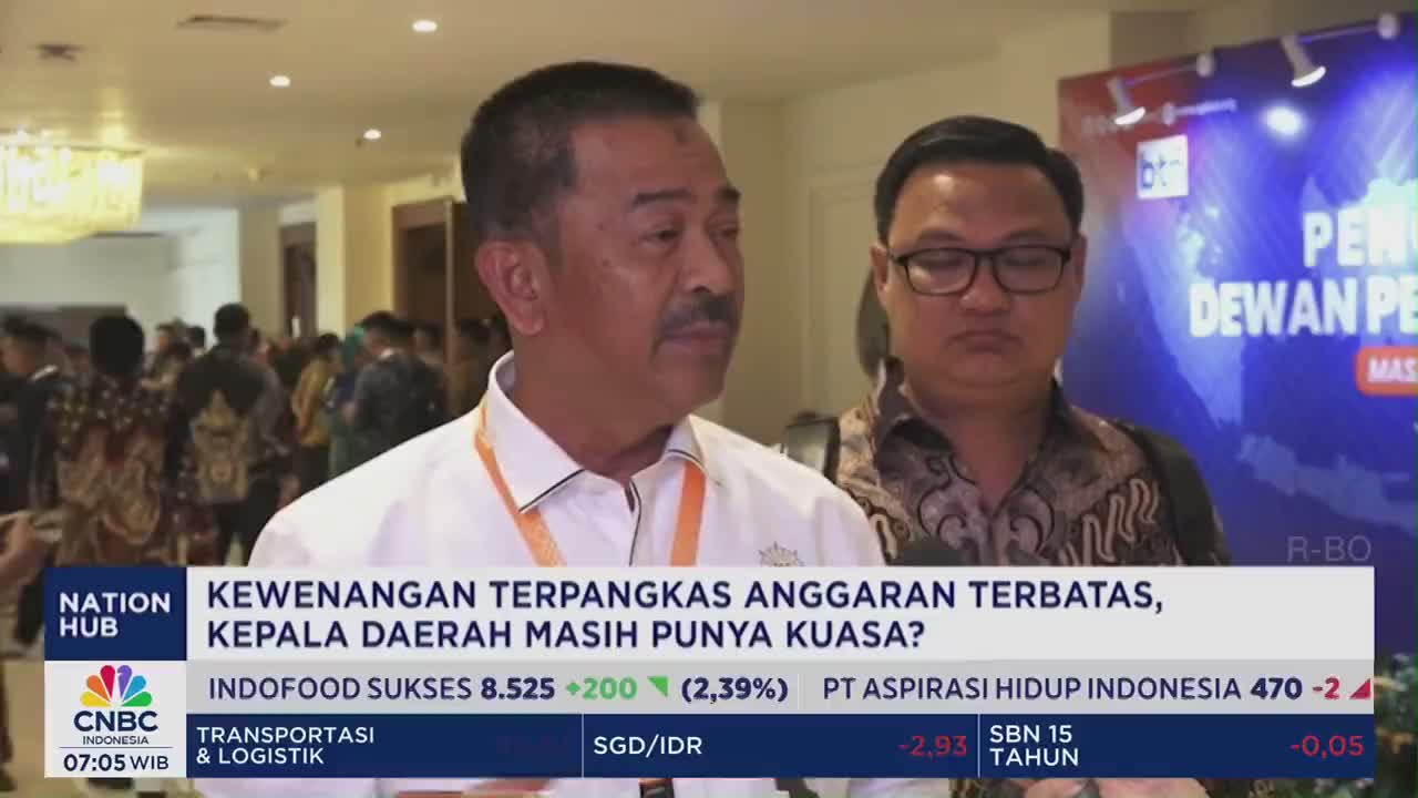 A man in a white shirt speaks directly into a microphone as another man in glasses stands beside him. Behind them, a CNBC Indonesia graphic displays financial data and the headline "KEWENANGAN TERPANGKAS ANGGARAN TERBATAS, KEPALA DAERAH MASIH PUNYA KUASA?"
