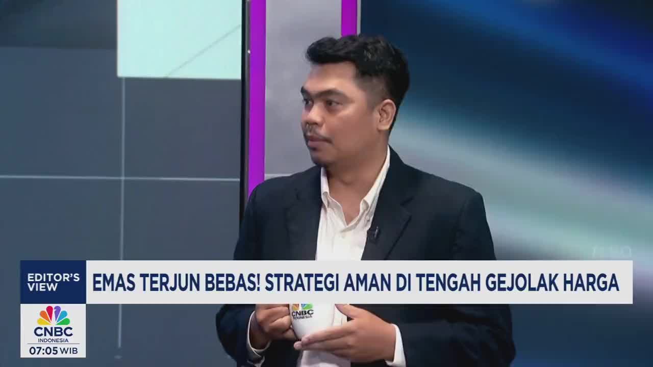 A man in a suit, holding a CNBC Indonesia mug, is looking to his right. The screen behind him displays the words "EMAS TERJUN BEBAS!"

