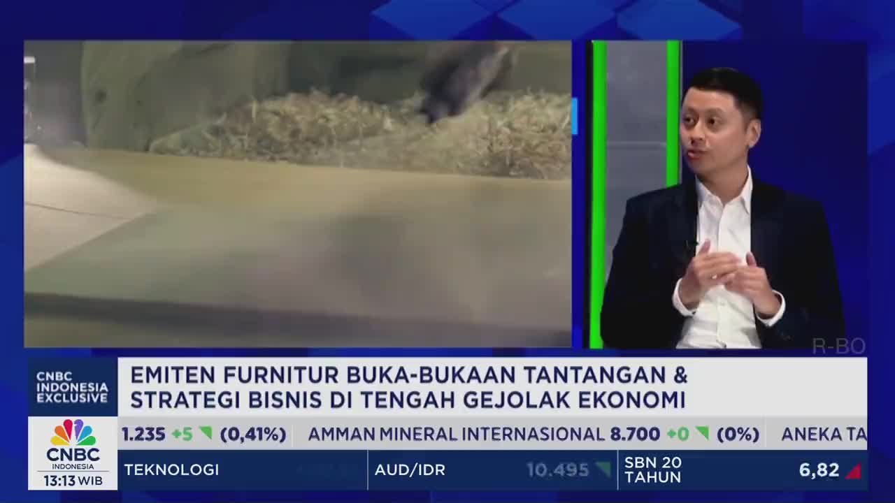A man in a suit gestures with his hands, speaking on a CNBC Indonesia program. The screen behind him shows a close-up of what looks like a wood-working process.
