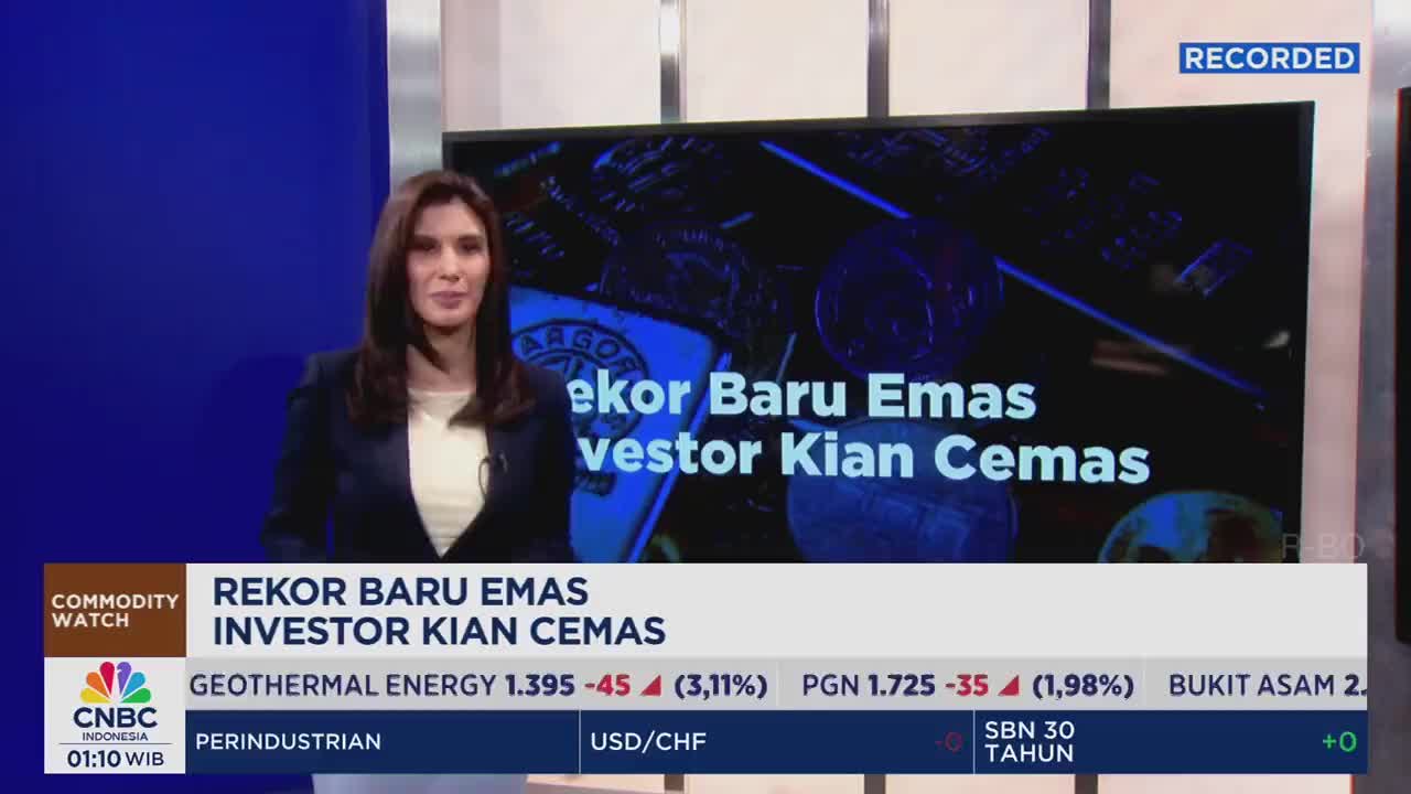 A woman in a blazer stands in front of a screen displaying the CNBC Indonesia logo and a headline about gold. Below, financial data scrolls, and the broadcast is marked as "recorded."
