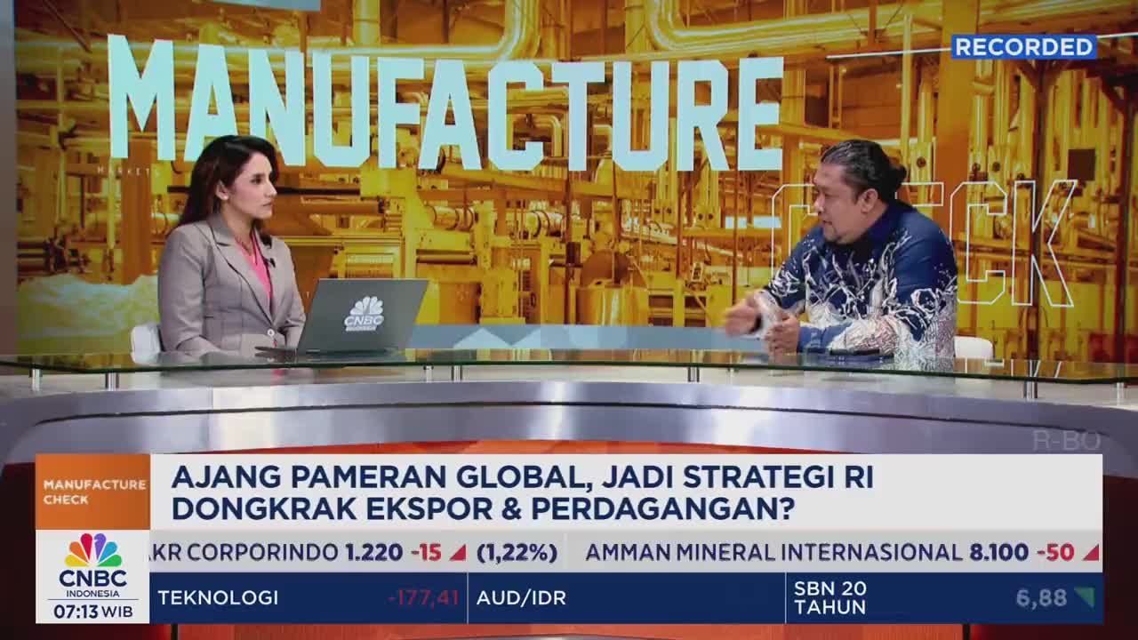 A woman in a blazer sits at a desk, looking at her laptop on the CNBC Indonesia set. Across from her, a man gestures while speaking, the backdrop displaying a manufacturing facility.
