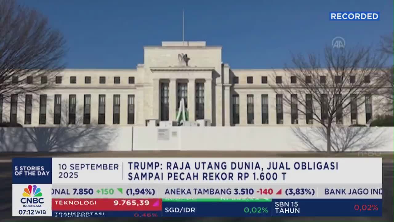 A stately building, likely the Federal Reserve, forms the backdrop for a CNBC Indonesia broadcast. The lower third of the screen displays financial data and a headline about Trump and debt.
