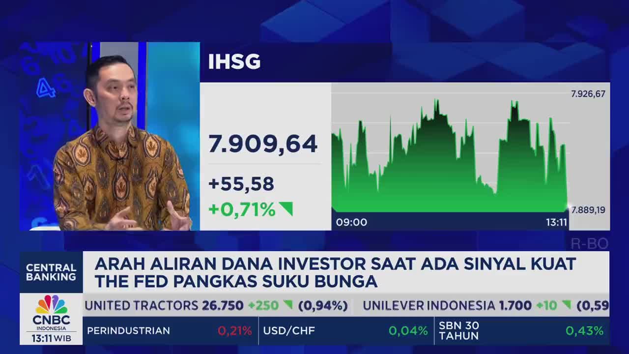 A man in a patterned shirt speaks on a CNBC Indonesia broadcast, gesturing with his hands. Behind him, a green graph charts the IHSG index, its peaks and valleys displayed on a screen.
