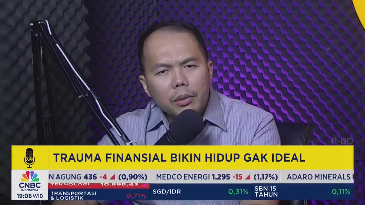 A man in a button-down shirt speaks into a microphone, framed by a yellow banner from CNBC Indonesia. The screen below him displays financial data and the headline "TRAUMA FINANSIAL BIKIN HIDUP GAK IDEAL."
